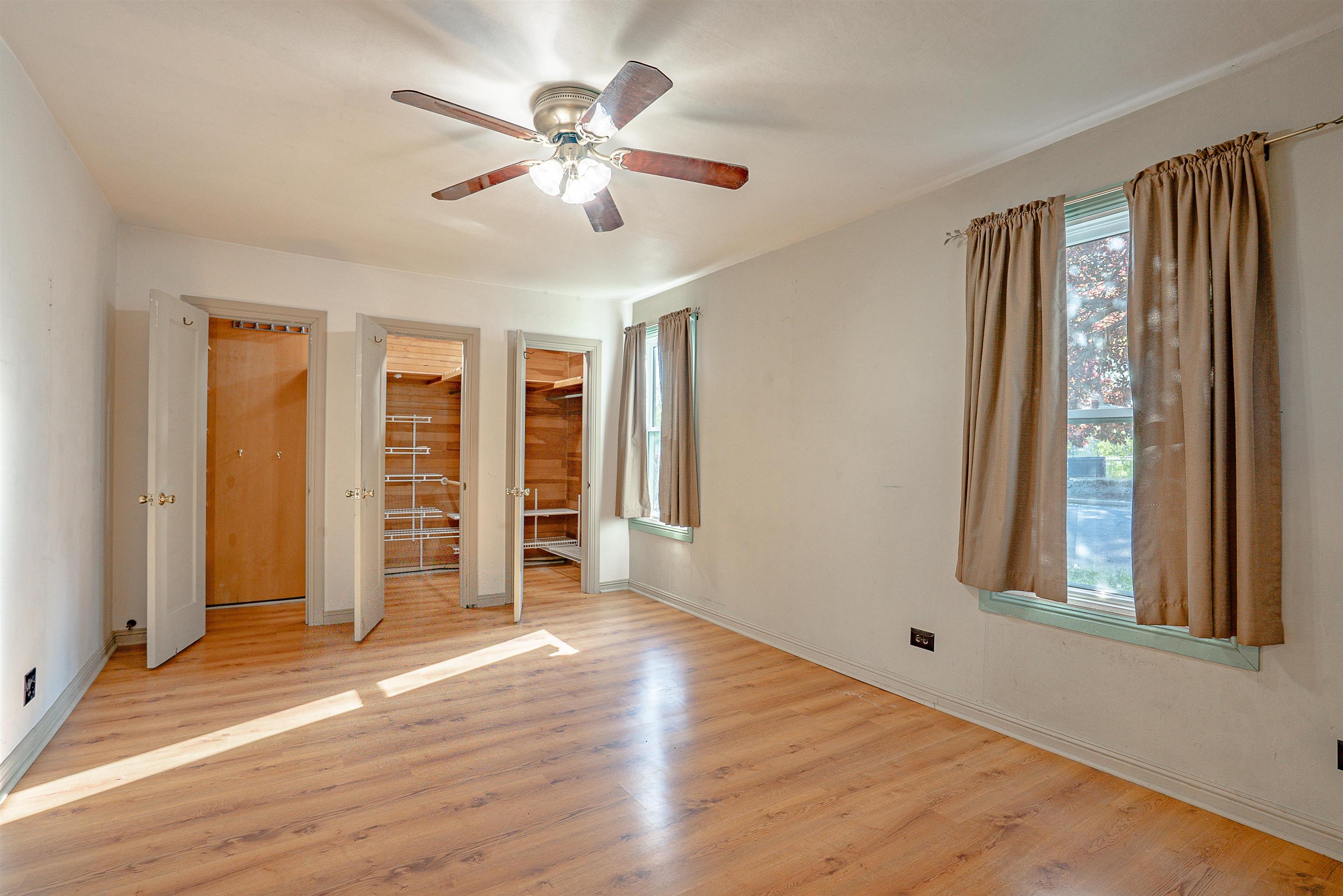 Empty bedroom with three open closets, wood flooring, curtained windows, and a ceiling fan.
