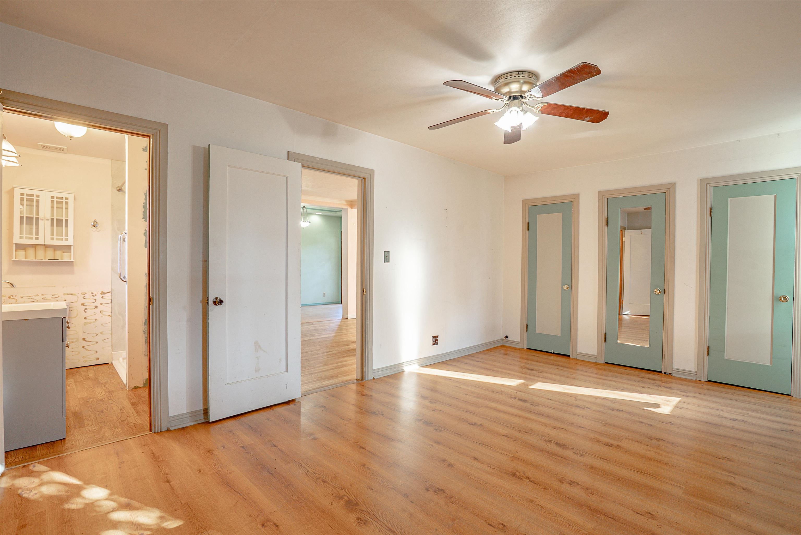 Empty bedroom with wood floors, a ceiling fan, and multiple doors, including an open bathroom doorway.