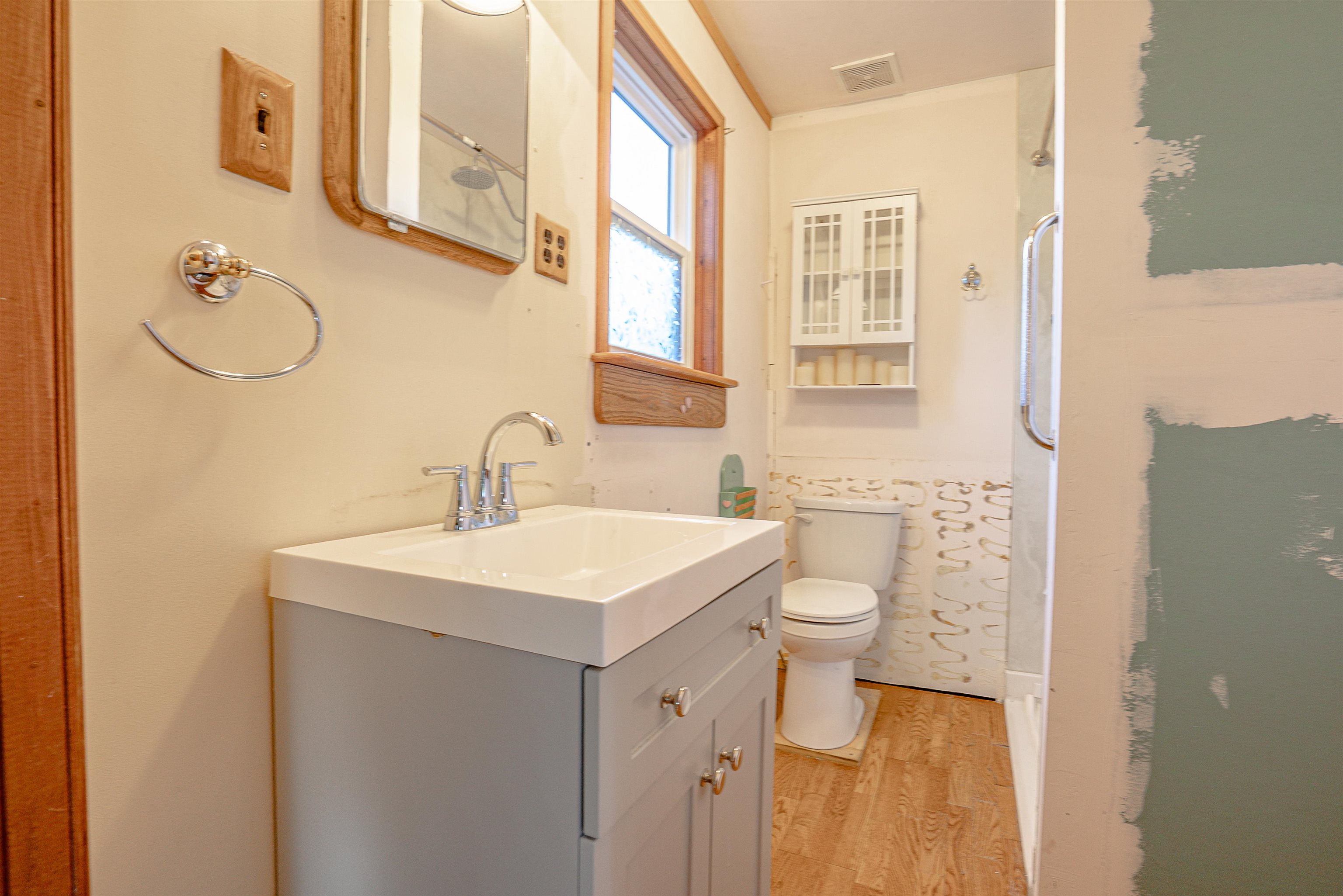 Bathroom with gray vanity, white sink, toilet, glass shower, and patchy paint on the wall.