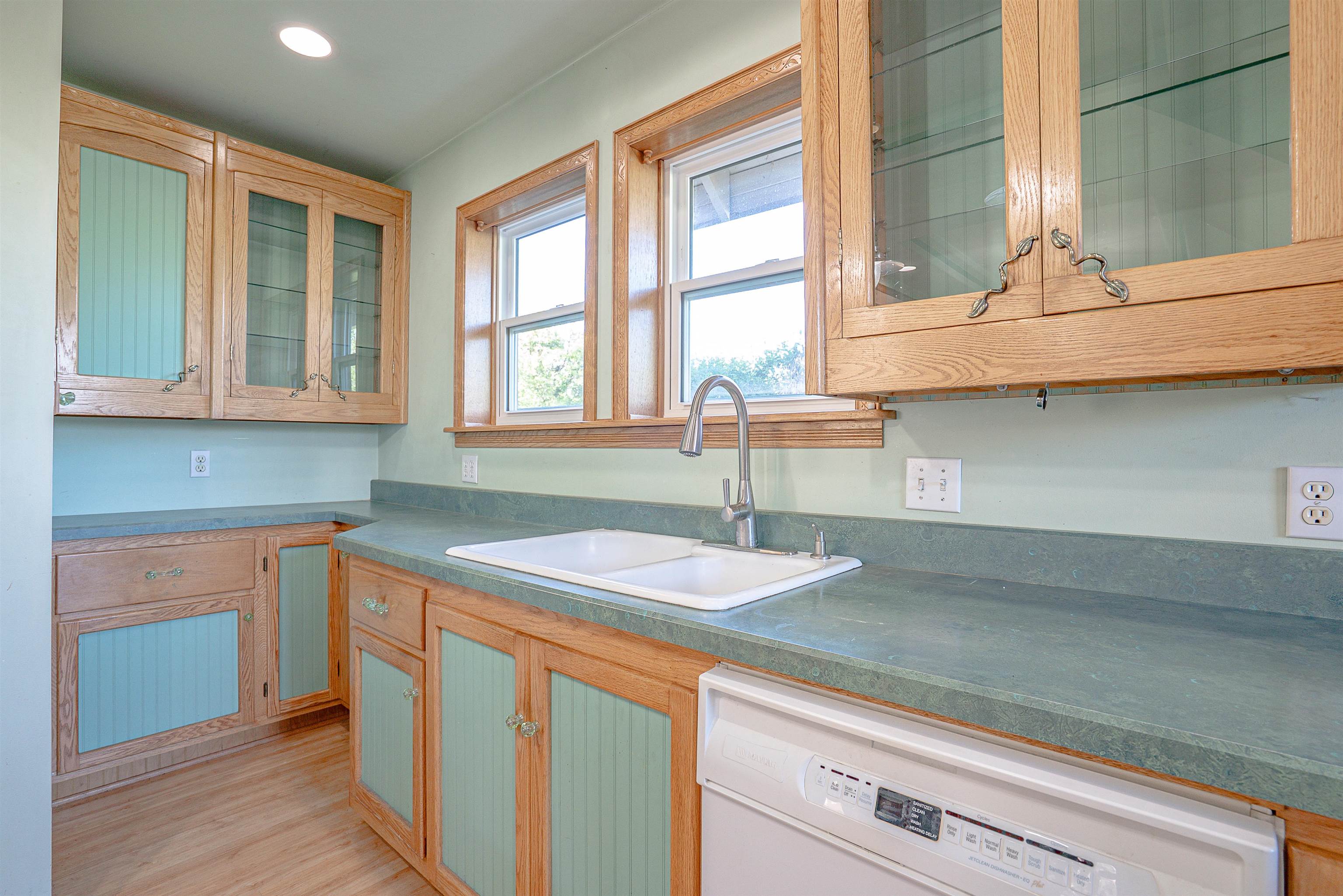 Kitchen sink beneath two windows, surrounded by green countertops and light wood cabinets.