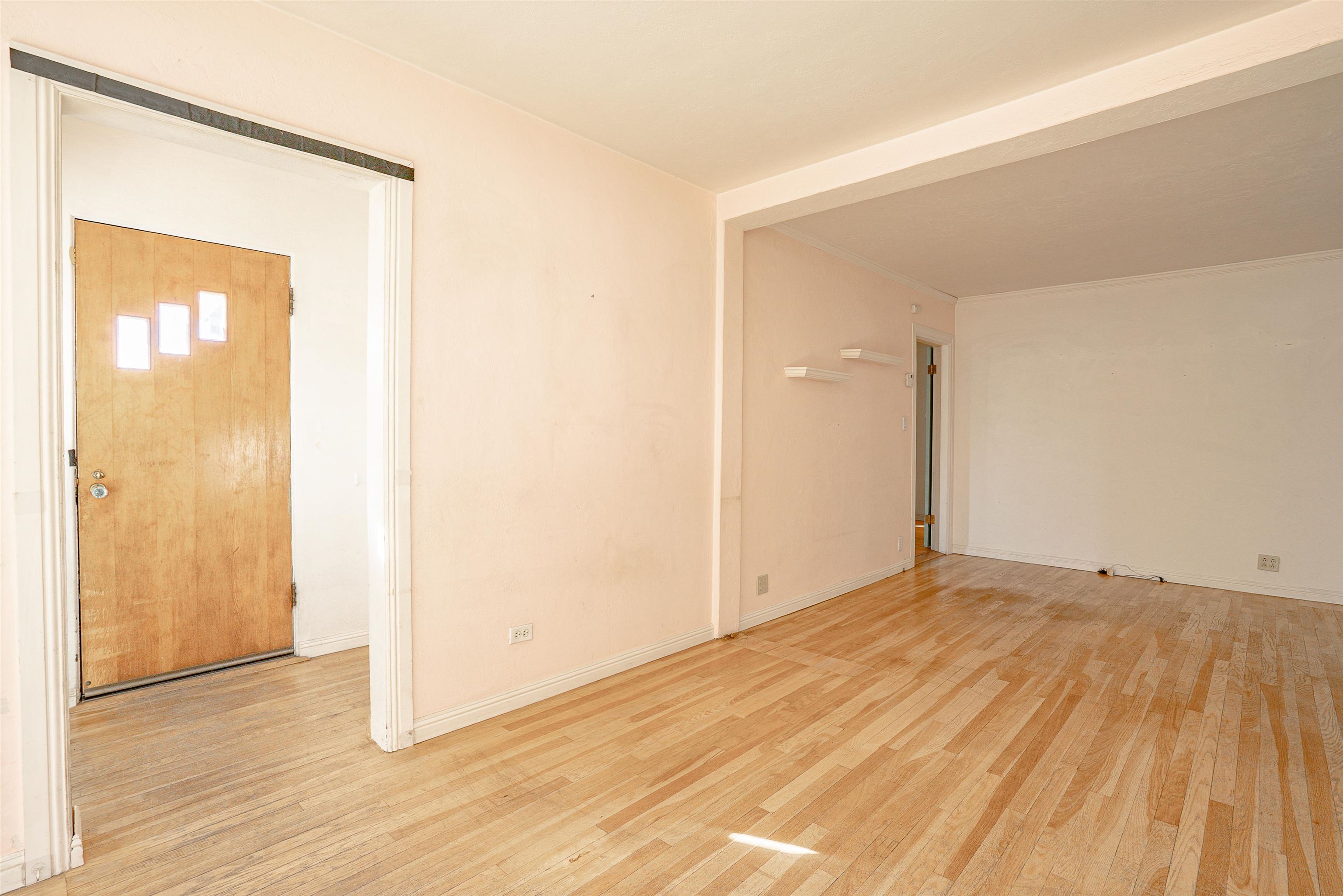 Empty living area with light hardwood floors, a wooden entry door, and two floating shelves.