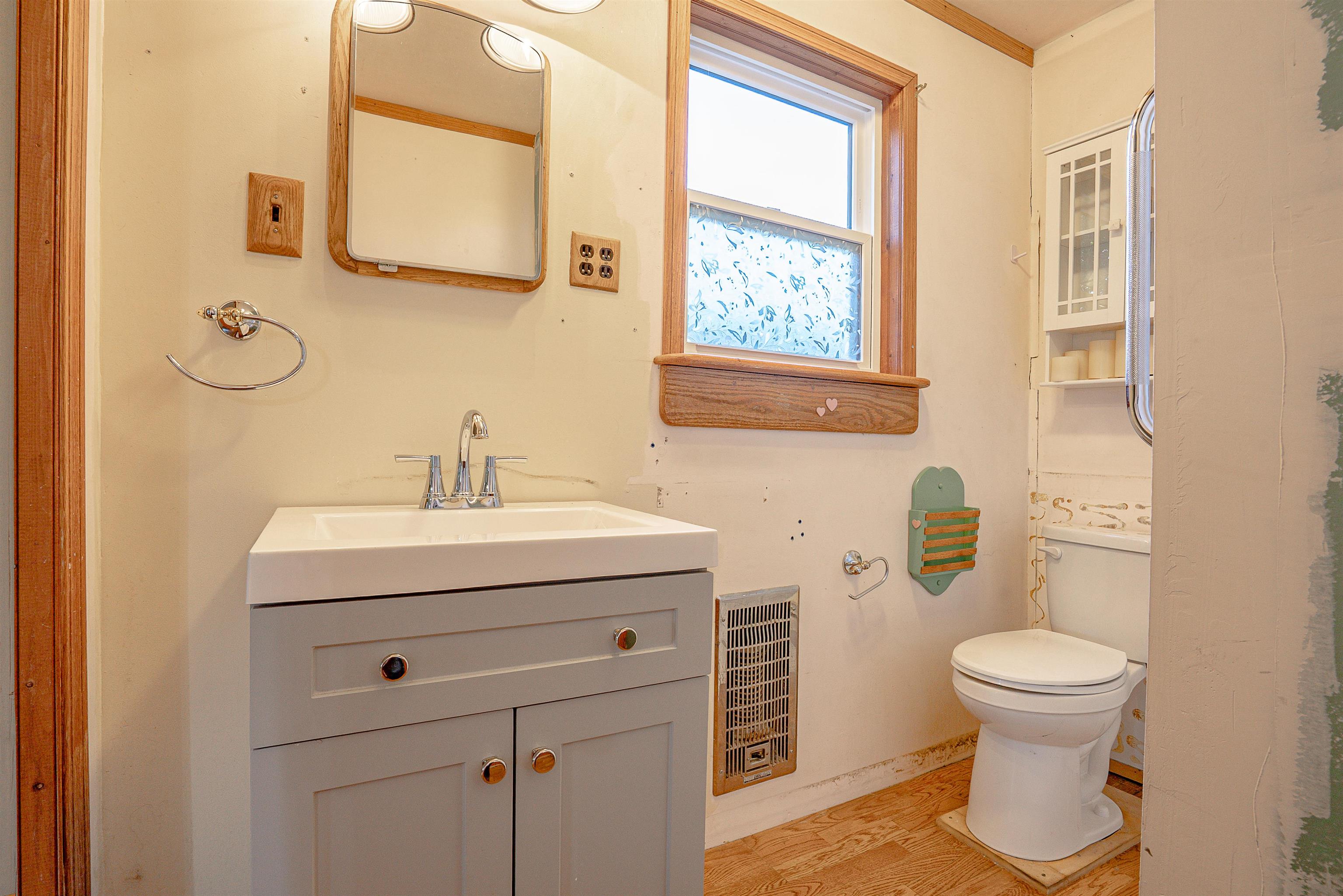 Bathroom with a gray vanity, wood-framed mirror and window, toilet, and wood flooring.