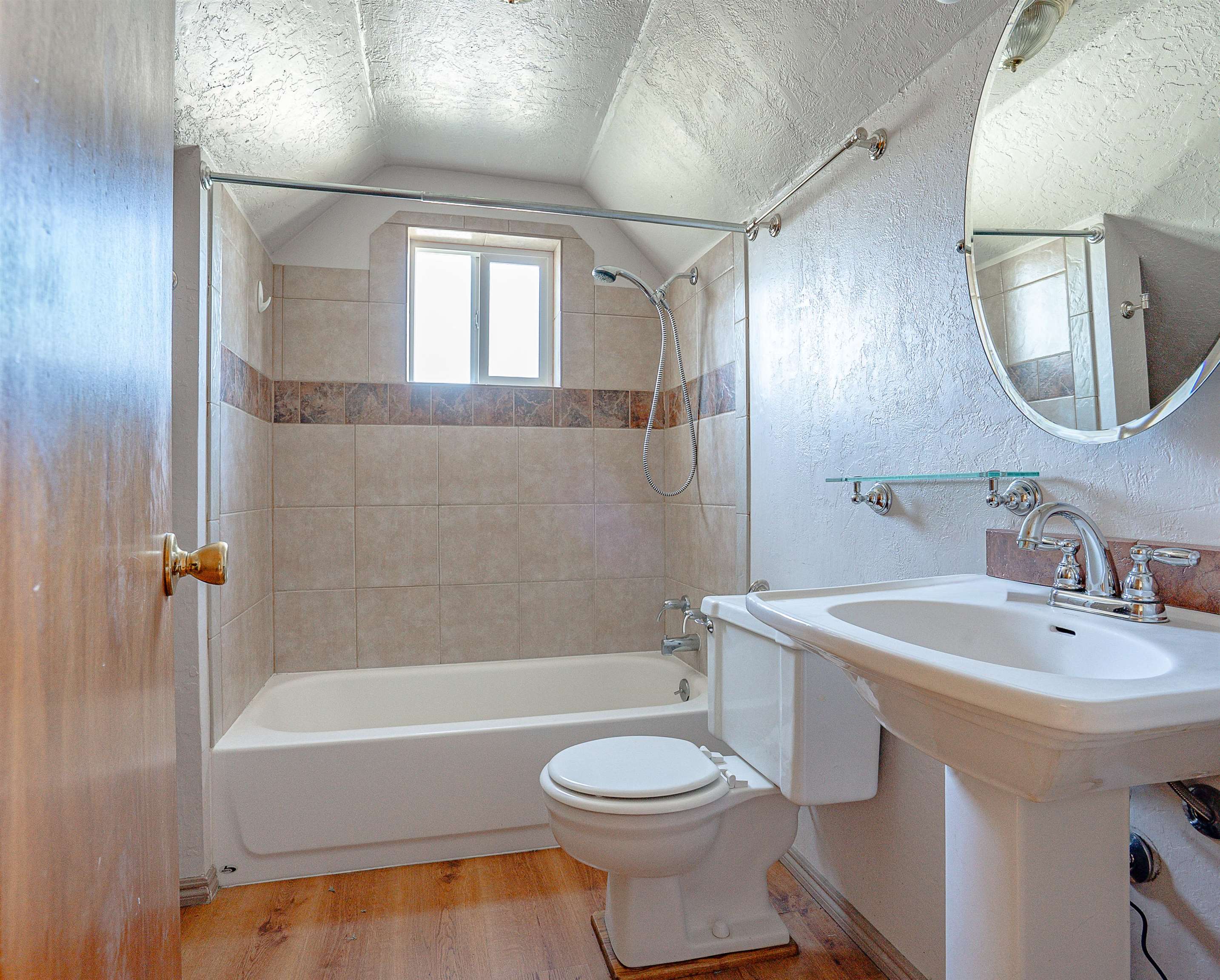 Bathroom with a tiled tub and shower, small window, pedestal sink, and wood-look floor.