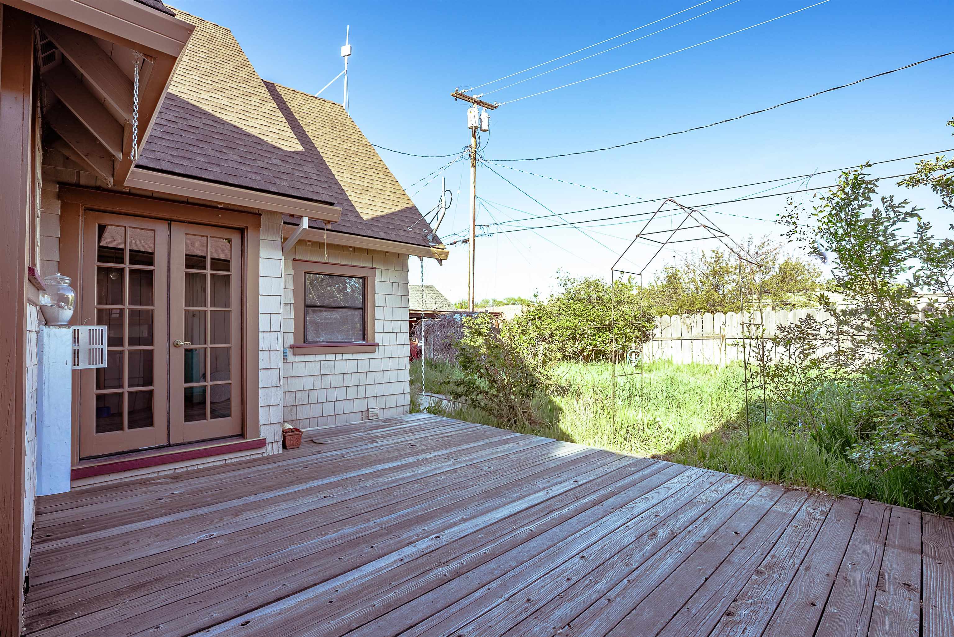Wooden backyard deck beside French doors, overlooking a fenced grassy yard with shrubs and a metal garden frame.