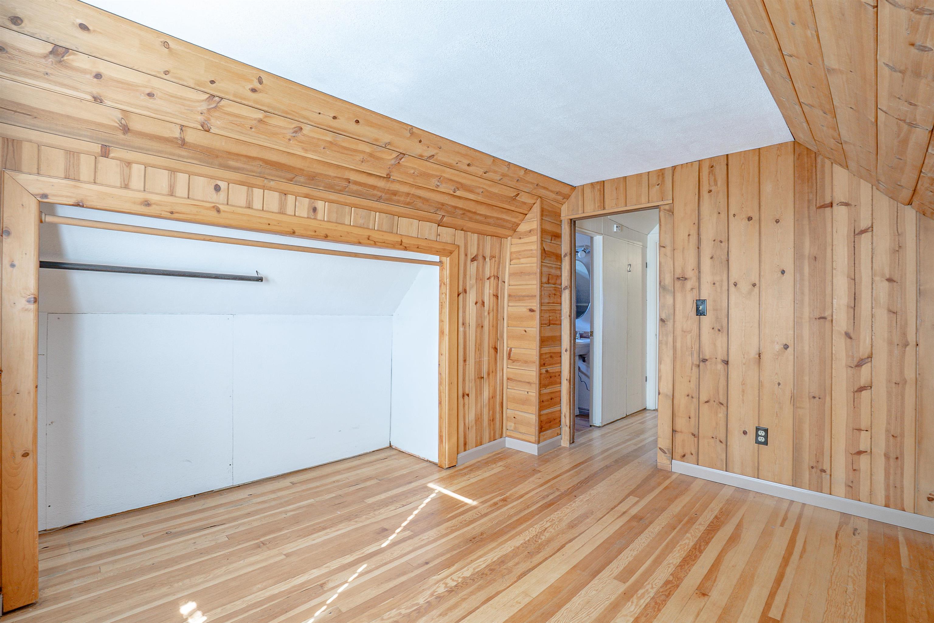 Empty bedroom with knotty pine walls, sloped wood ceiling, hardwood floors, and an open closet.