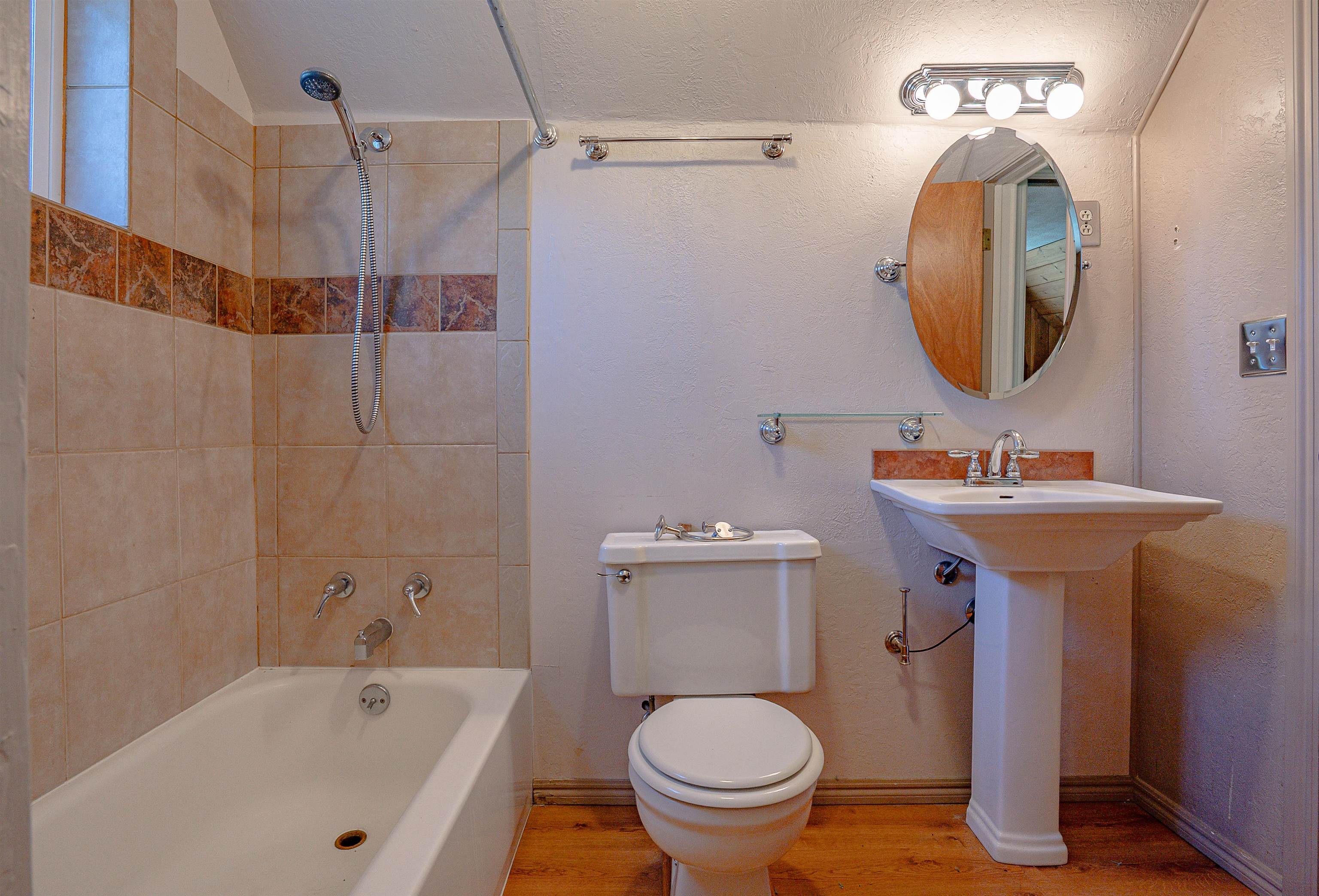 Bathroom with tiled tub and shower, toilet, pedestal sink, and oval mirror.