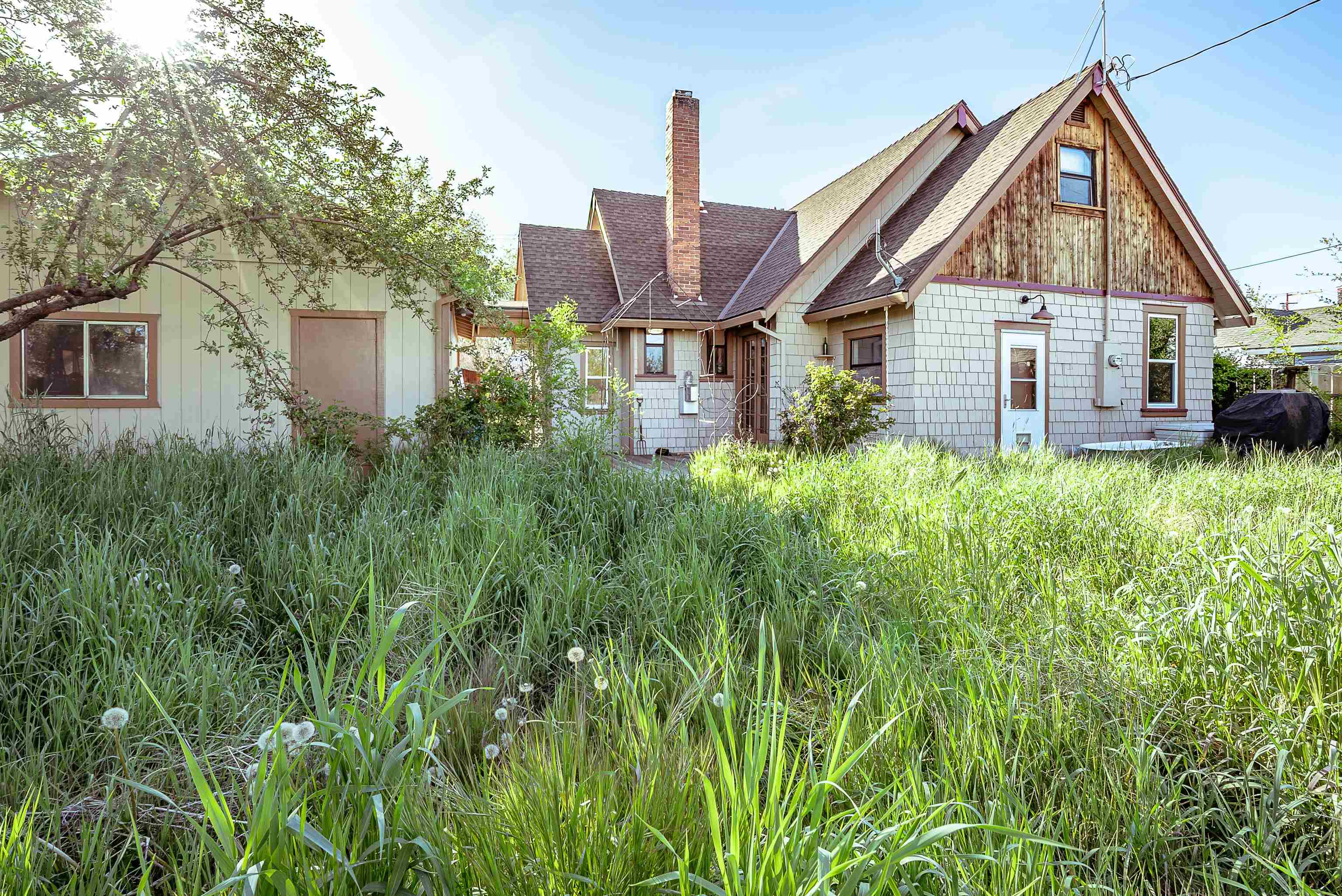 Multi-gabled house with gray siding and brick chimney behind tall, overgrown grass.