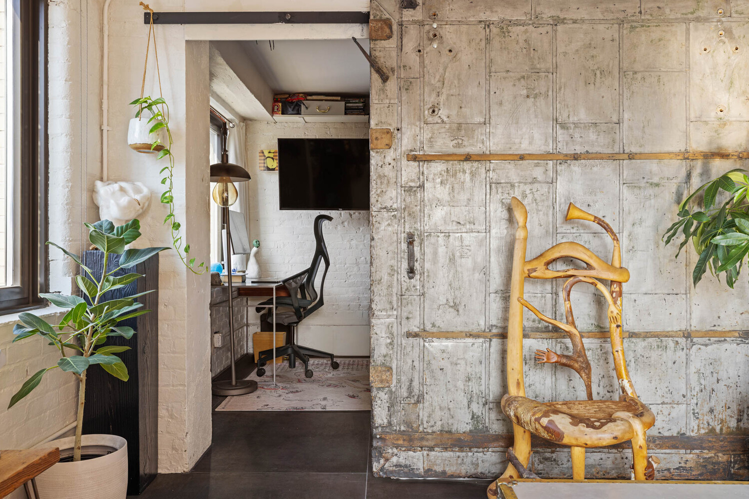 Home office nook behind a weathered sliding door, with a sculptural wooden chair and potted plants.