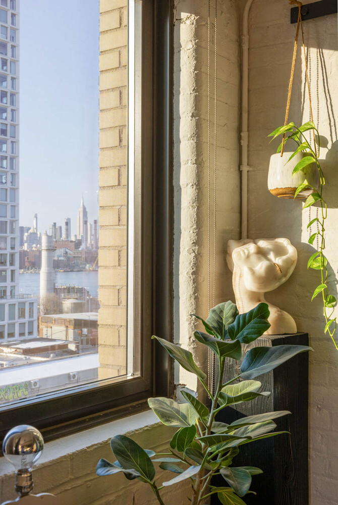 Window nook with plants and a sculptural bust overlooking the Manhattan skyline and water.