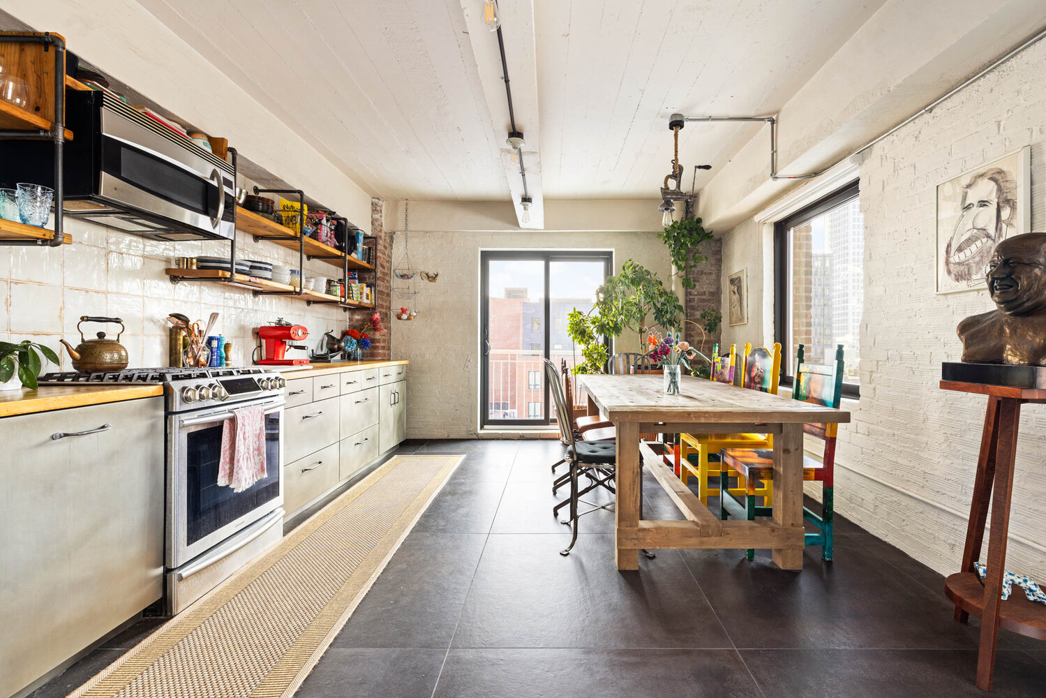 Loft kitchen and dining area with open shelves, a rustic table, colorful chairs, and large windows.