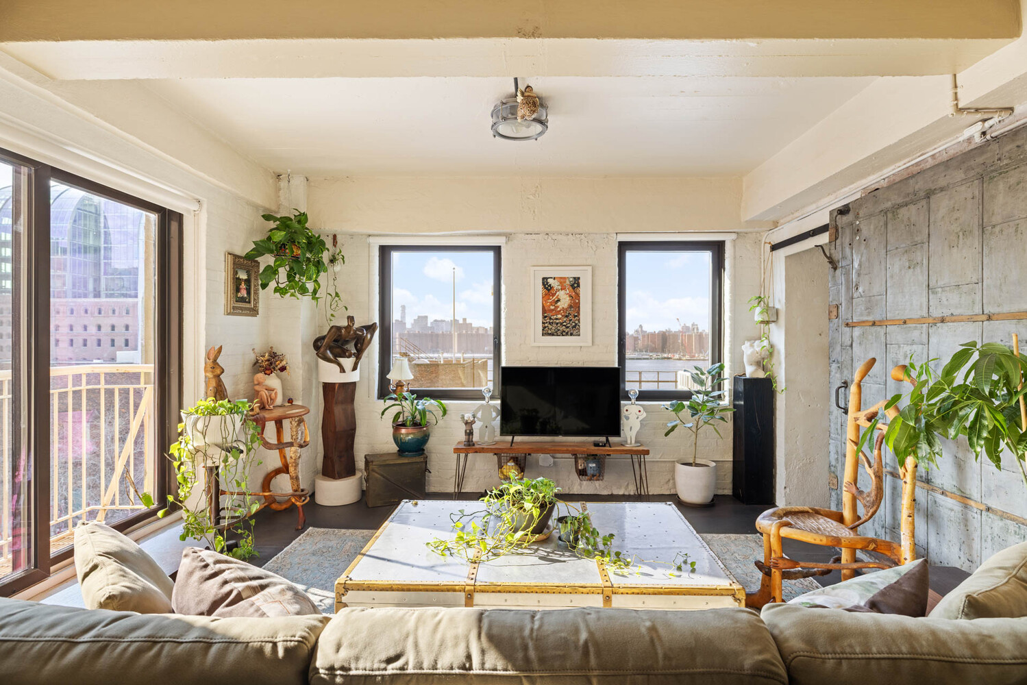 Living room with white brick walls, abundant plants, and windows overlooking the waterfront and city skyline.