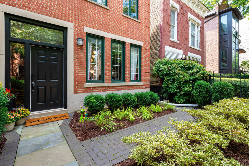 Red-brick home entrance with a black door, landscaped flower beds, and a paved walkway.