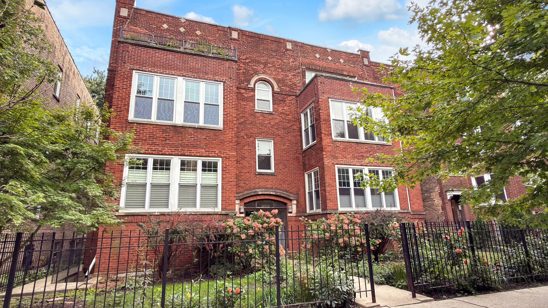 Red brick residential building with white-framed windows, a fenced garden, and flowering shrubs.