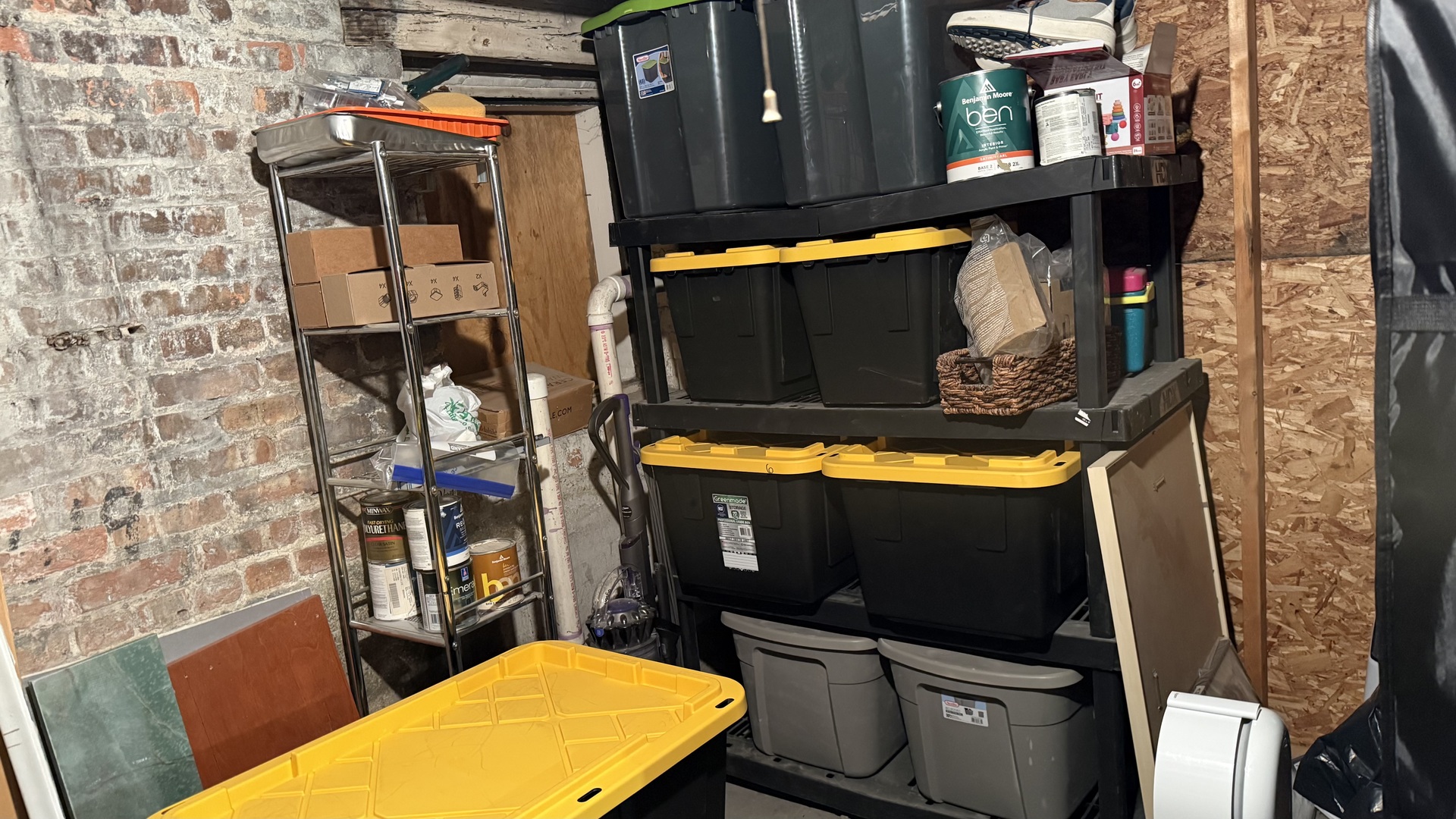 Basement storage area with stacked plastic bins, paint cans, and boxes beside exposed brick walls.