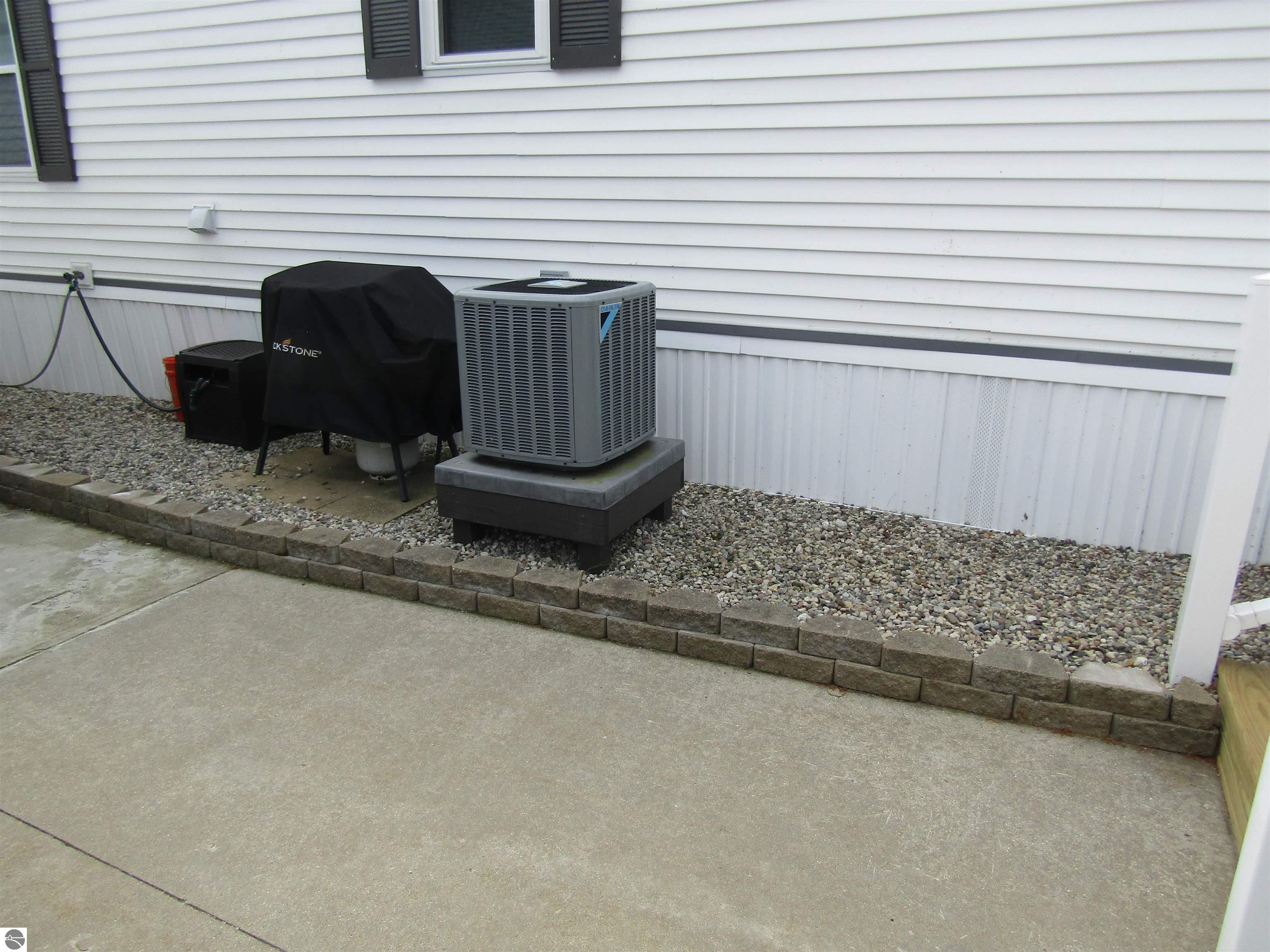 Outdoor air-conditioning condenser beside white house siding, set on a raised pad next to a gravel bed.