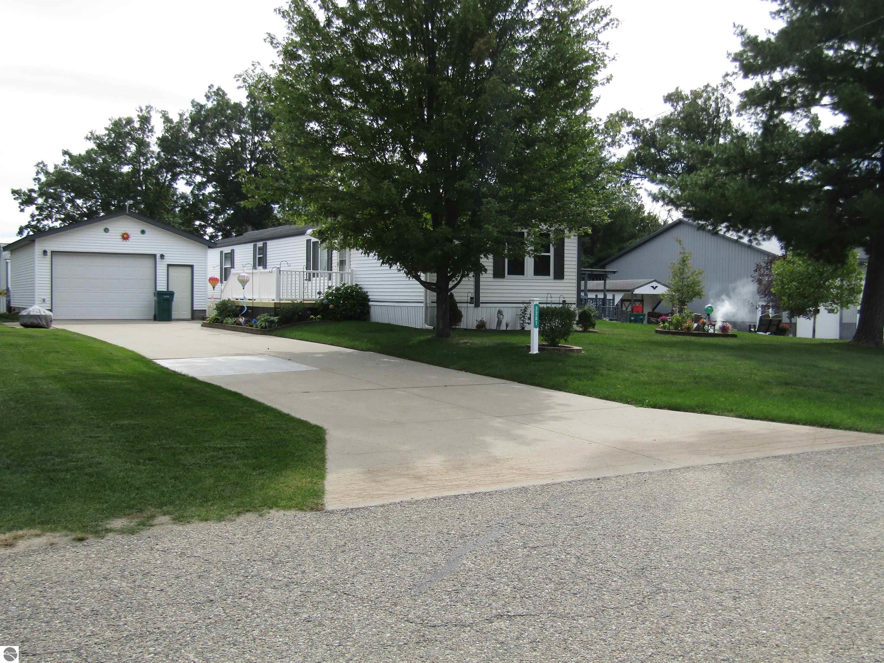 White single-story home with an attached garage and porch beneath mature trees beside a gravel road.