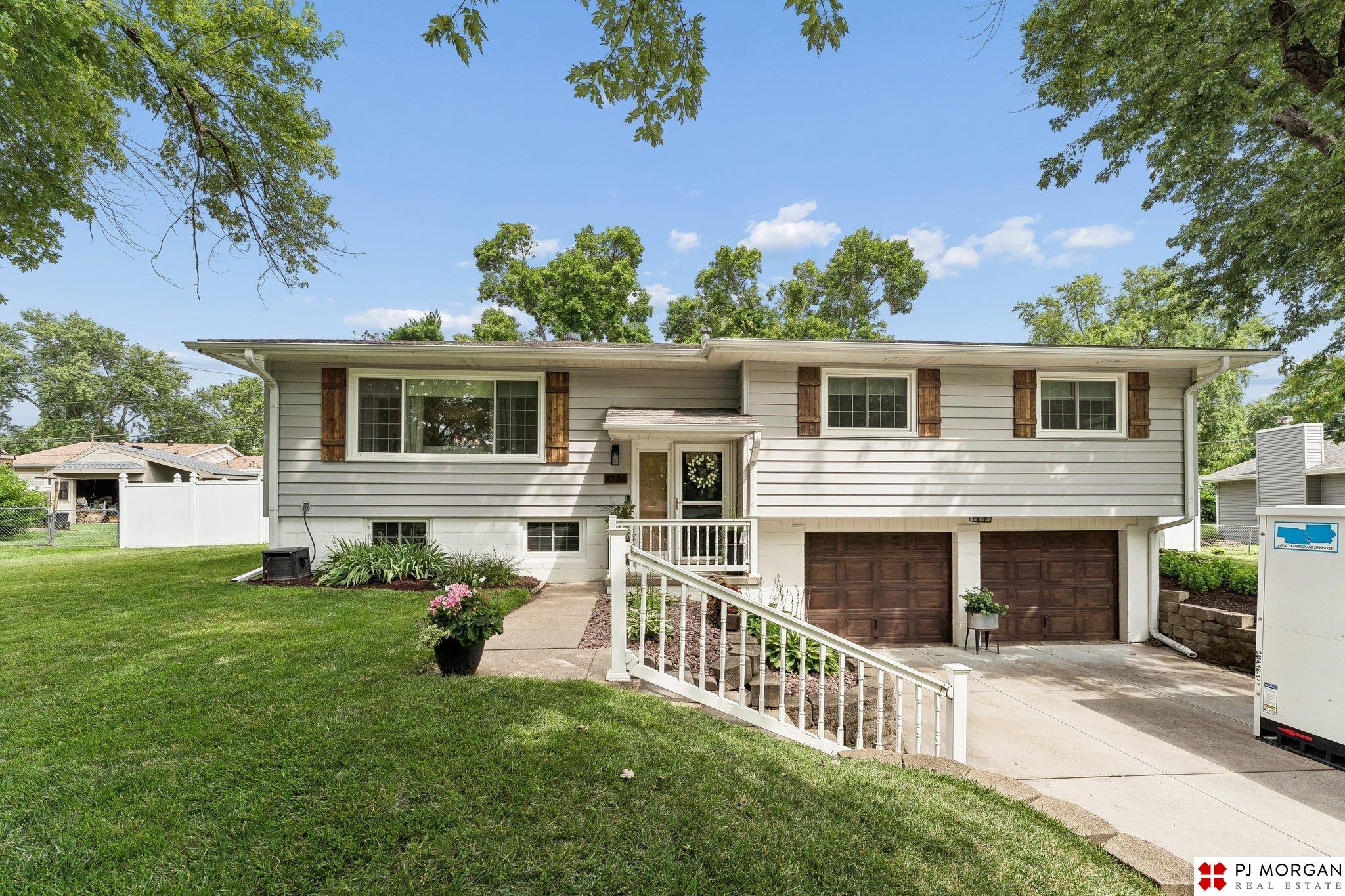 White split-level home with brown shutters, double garage, sloping entry walkway, and a tree-shaded lawn.