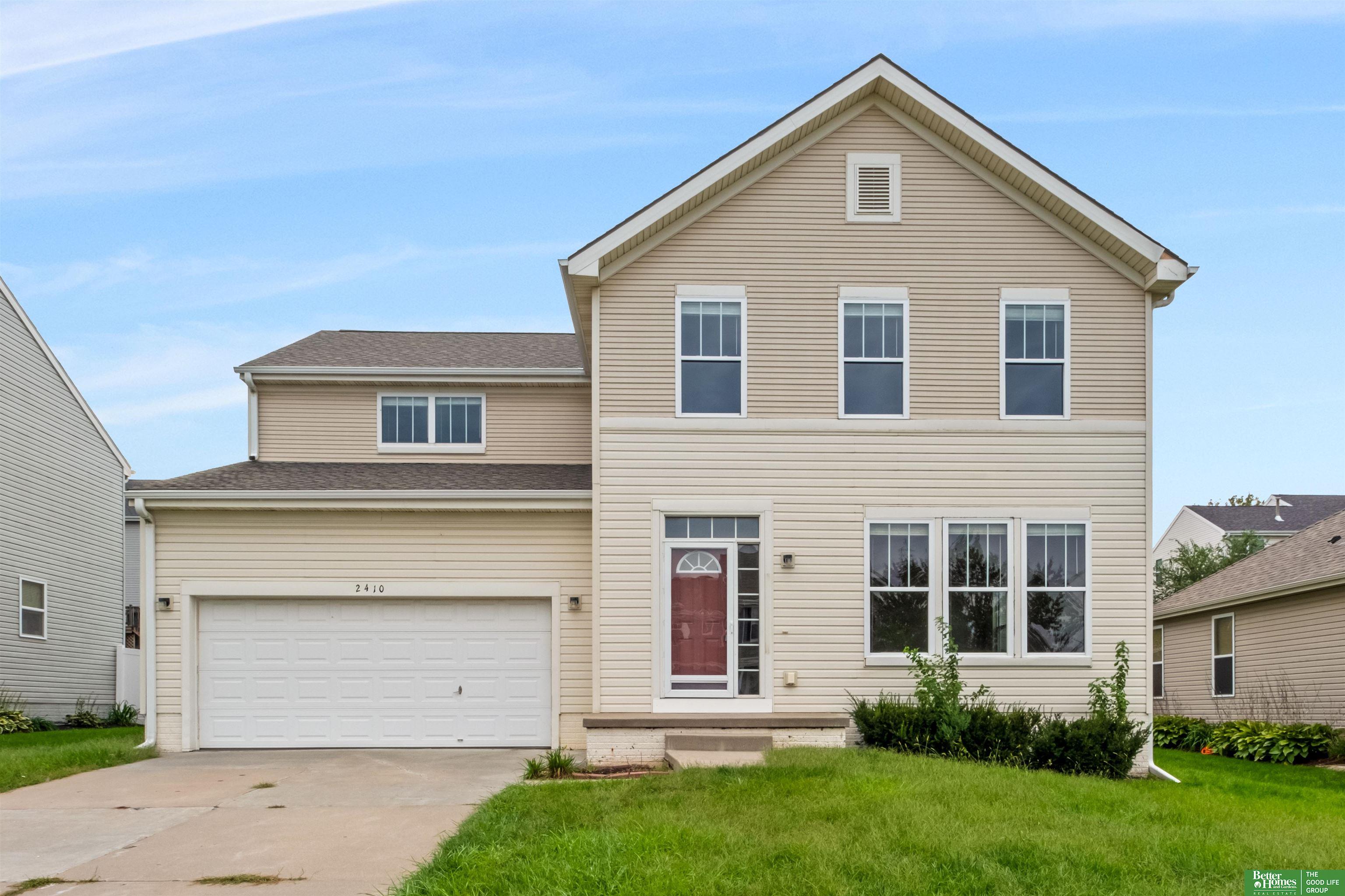 Two-story beige-sided home with a two-car garage, red front door, and broad green lawn.