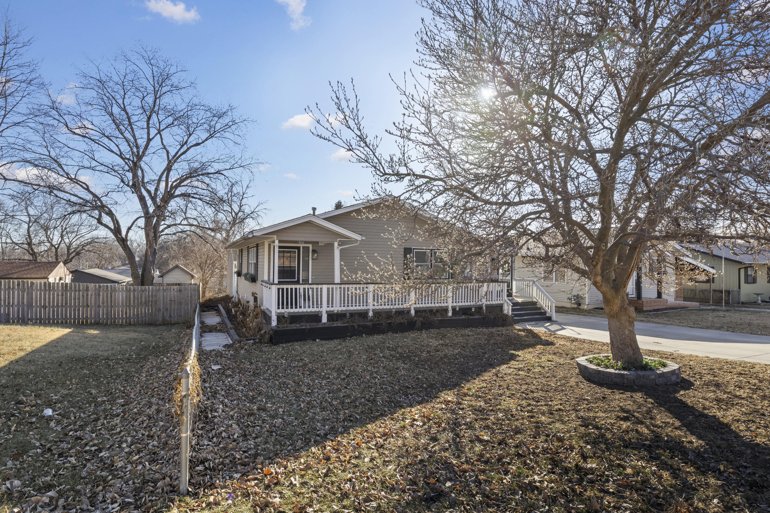 Gray house with a raised white-railed porch, fenced yard, and large leafless tree.