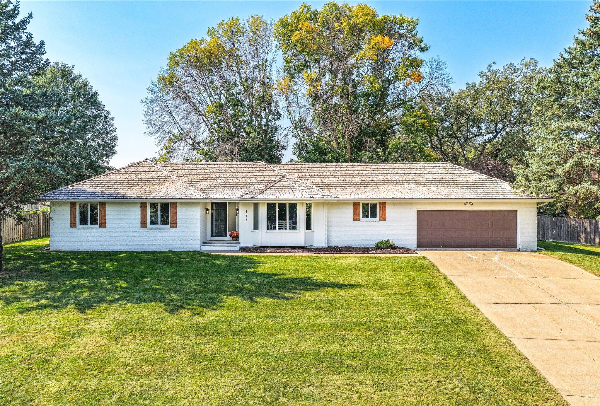 Single-story white brick home with a two-car garage, broad shingle roof, and mature trees.