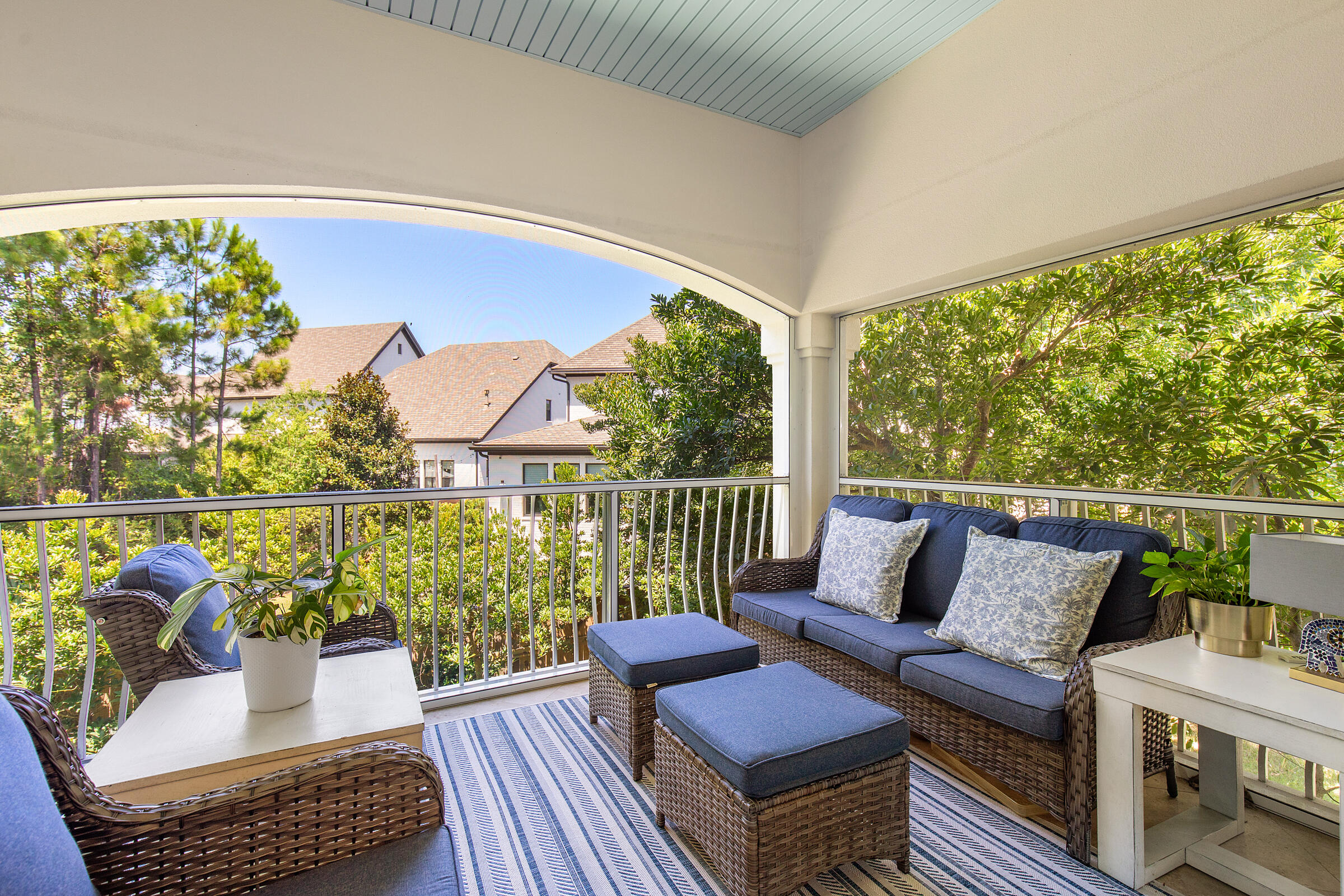 Covered patio furnished with wicker seating, blue cushions, and a striped rug overlooking leafy trees.