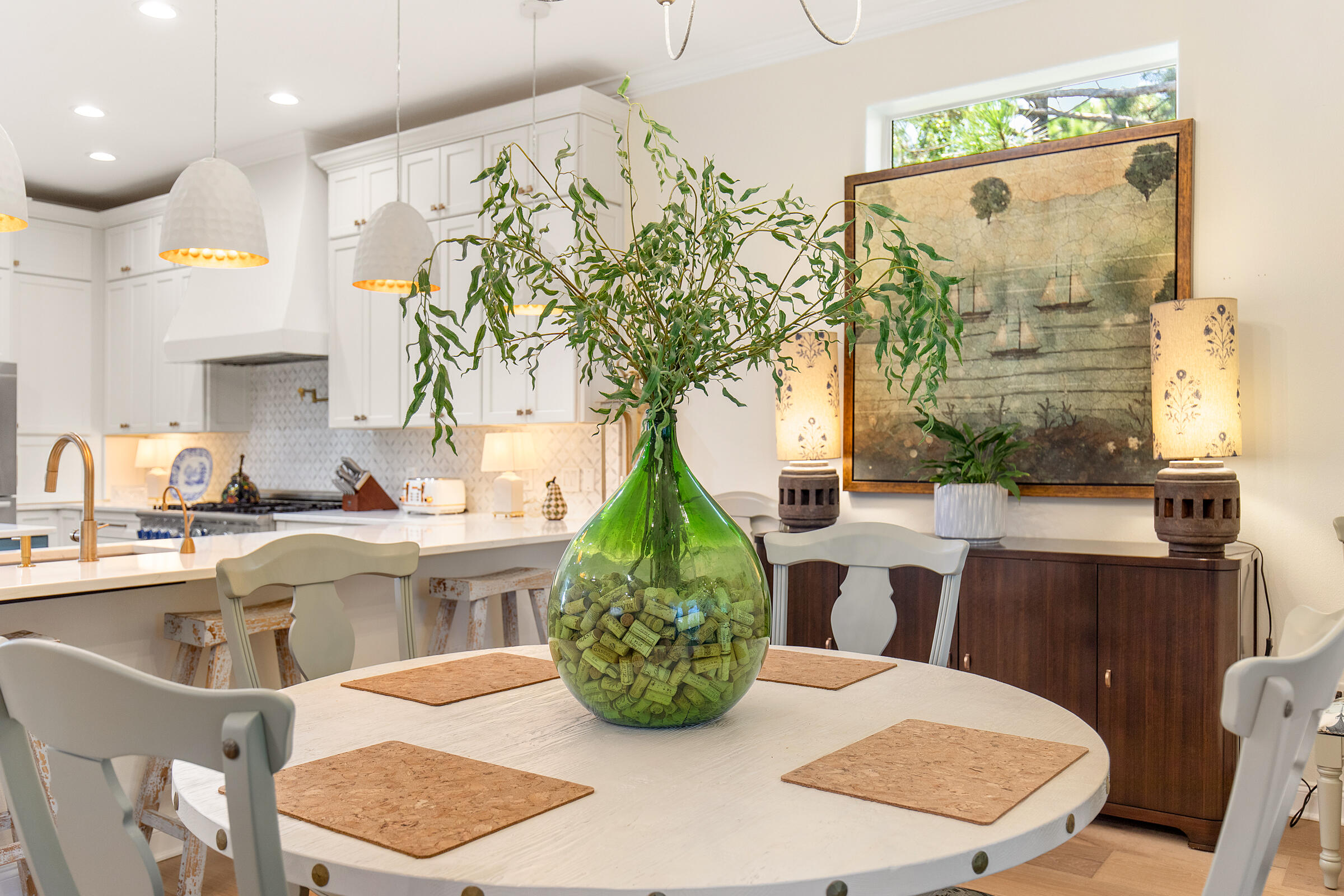 Dining area with a large white table and green vase beside a bright kitchen with white cabinetry.
