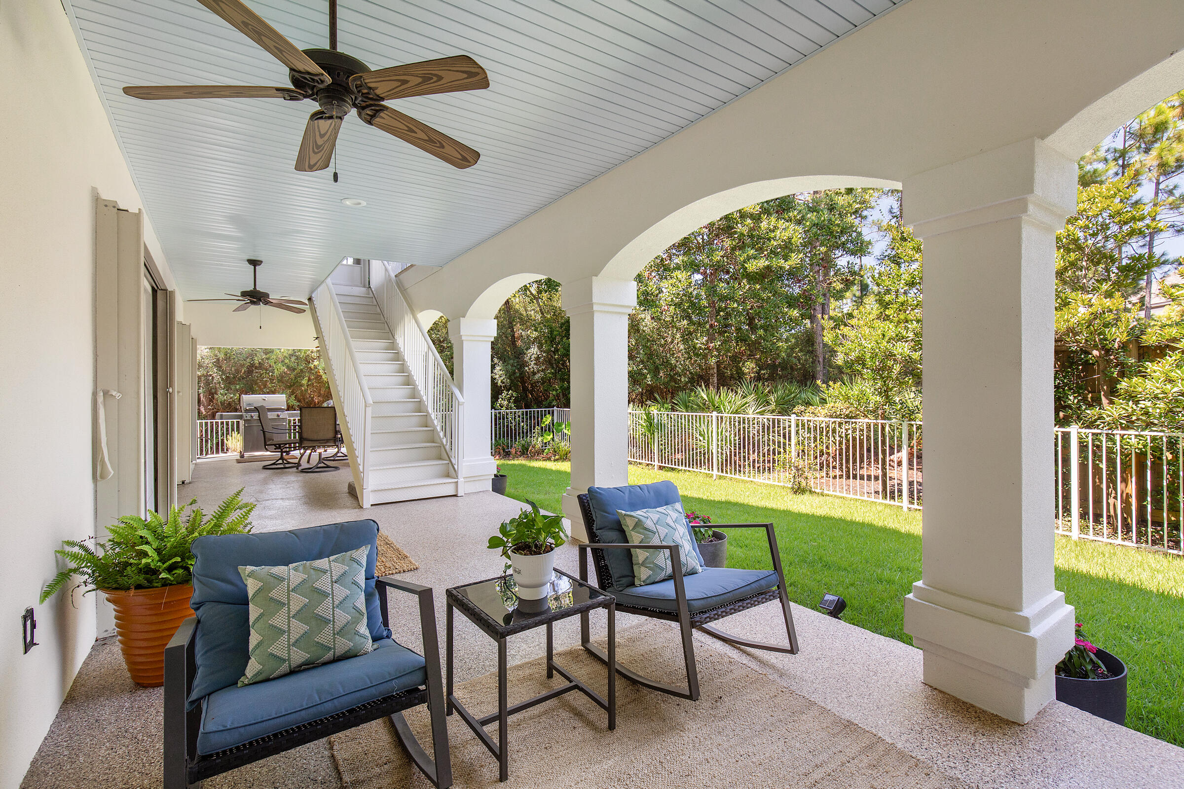 Covered patio with blue seating, white arches, and stairs overlooking a fenced lawn.
