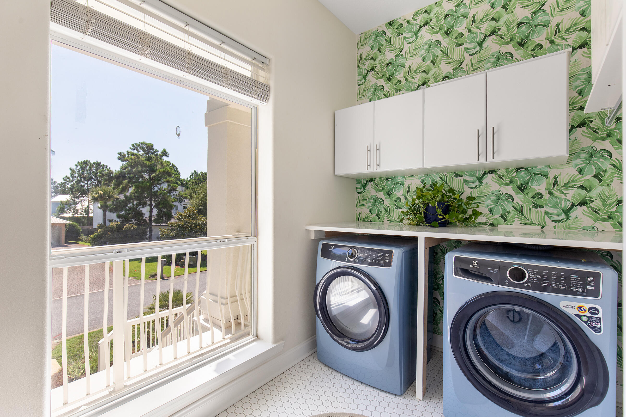 Laundry room with blue front-loading washer and dryer, botanical wallpaper, and a large window.