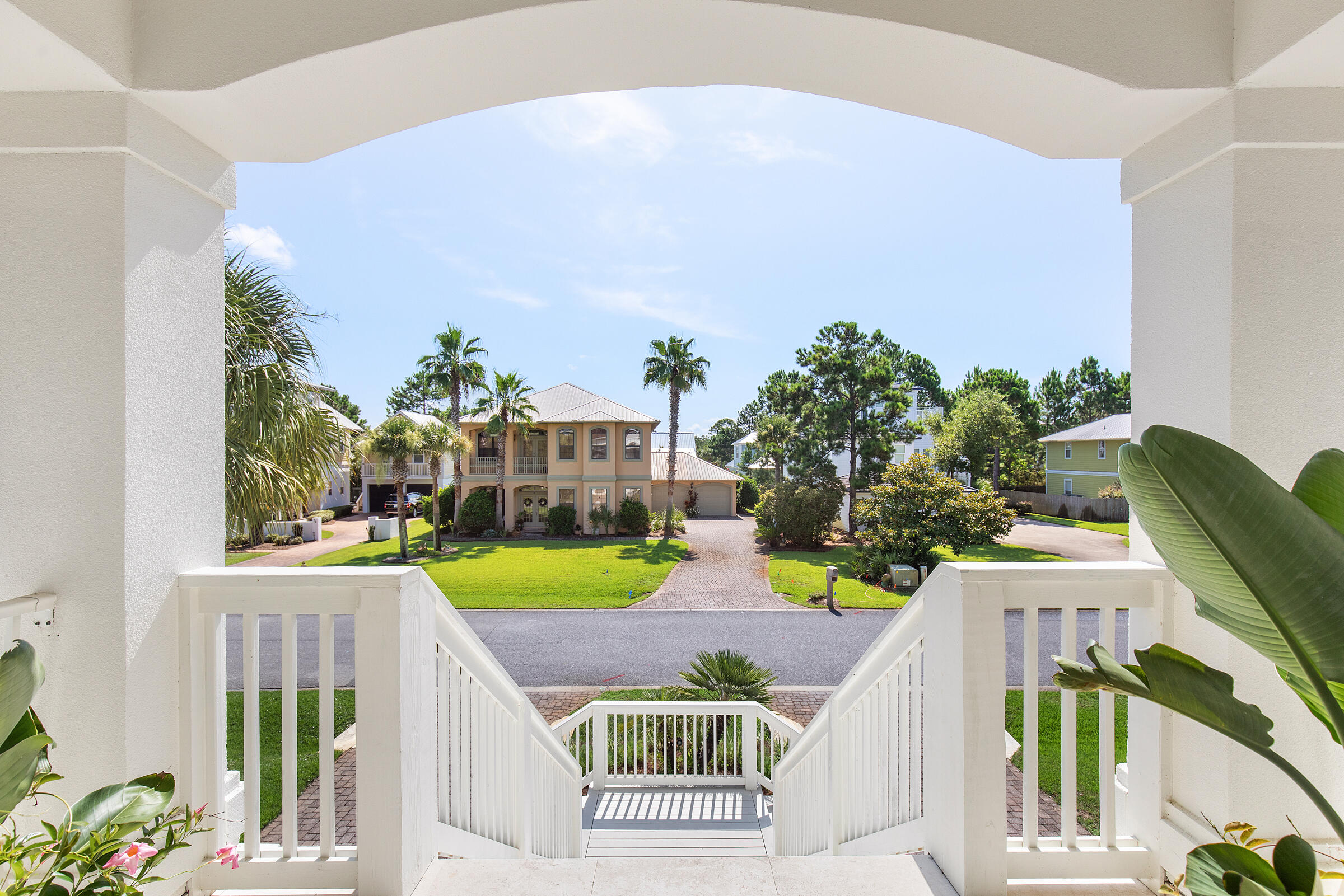 Covered porch stairs frame a two-story tan house across the street, with palm trees and a brick driveway.
