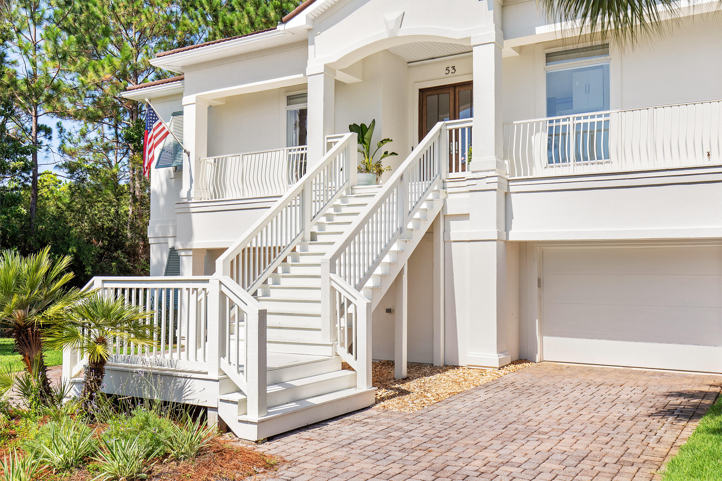 White home exterior with a wide staircase to the covered entry, balconies, garage, and tropical landscaping.