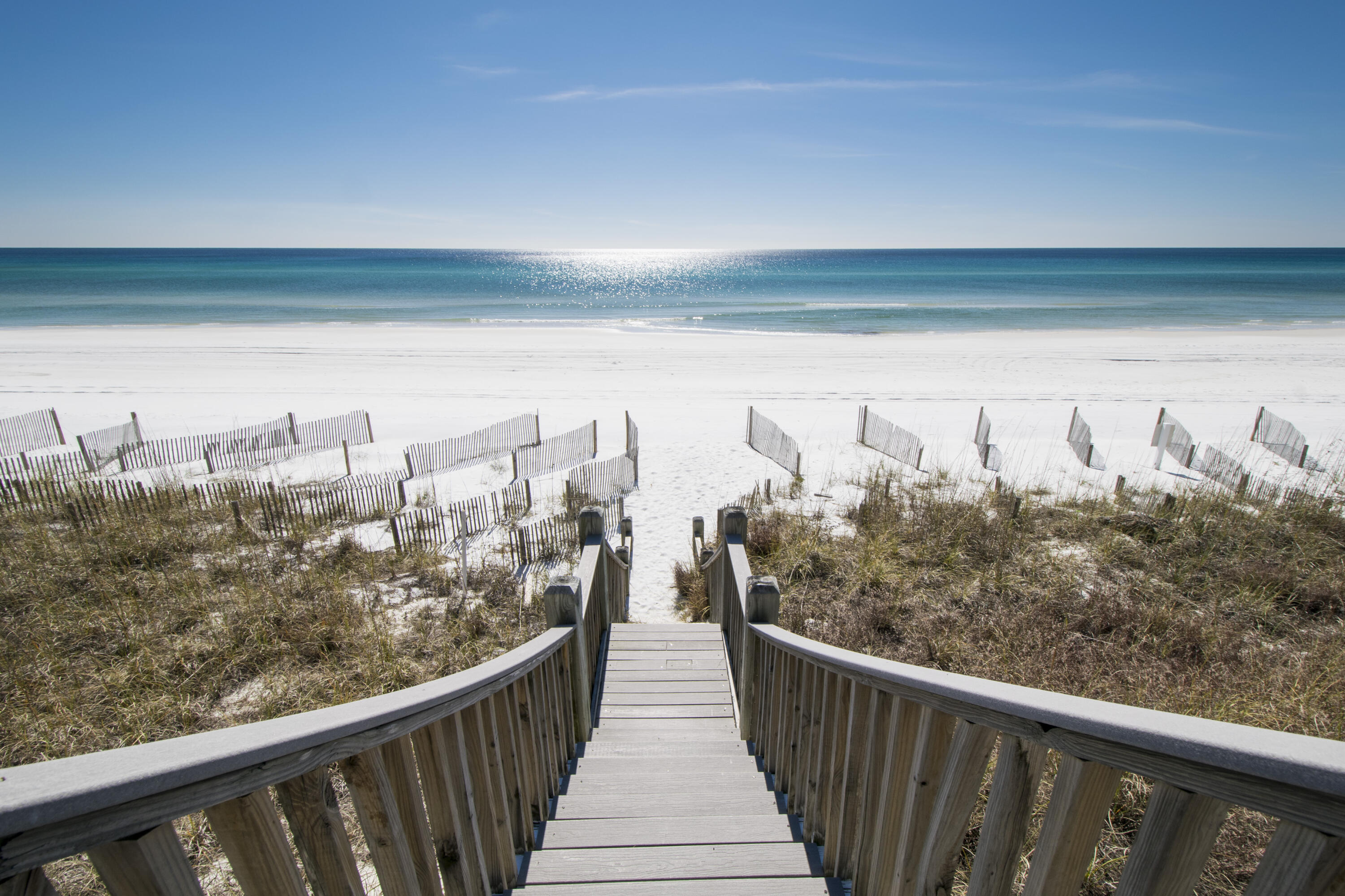 Beach access boardwalk crosses dune grass and white sand toward turquoise water.