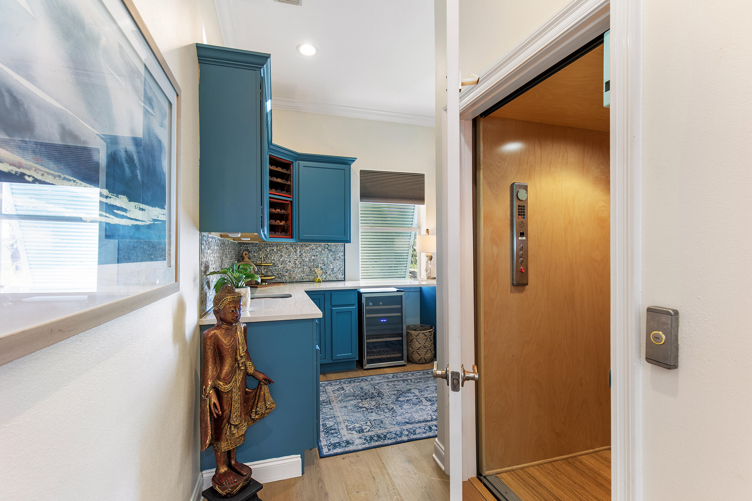Blue-cabinet wet bar with wine refrigerator beside an open wood-paneled elevator.