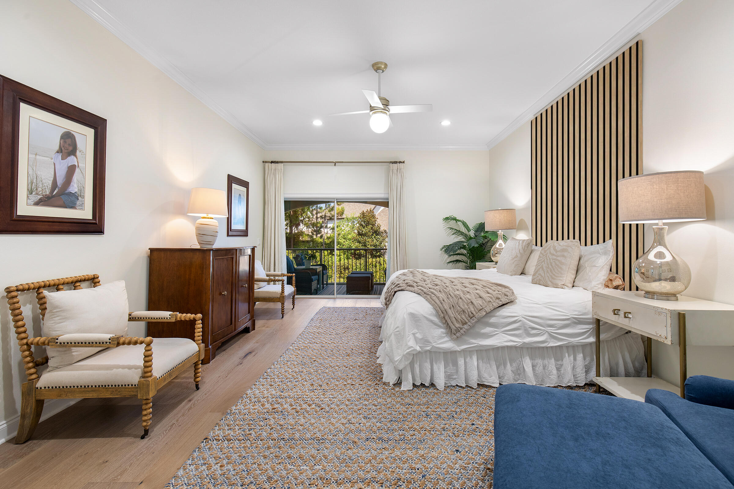 Furnished bedroom with a white bed, slatted accent wall, and sliding doors to a leafy balcony.