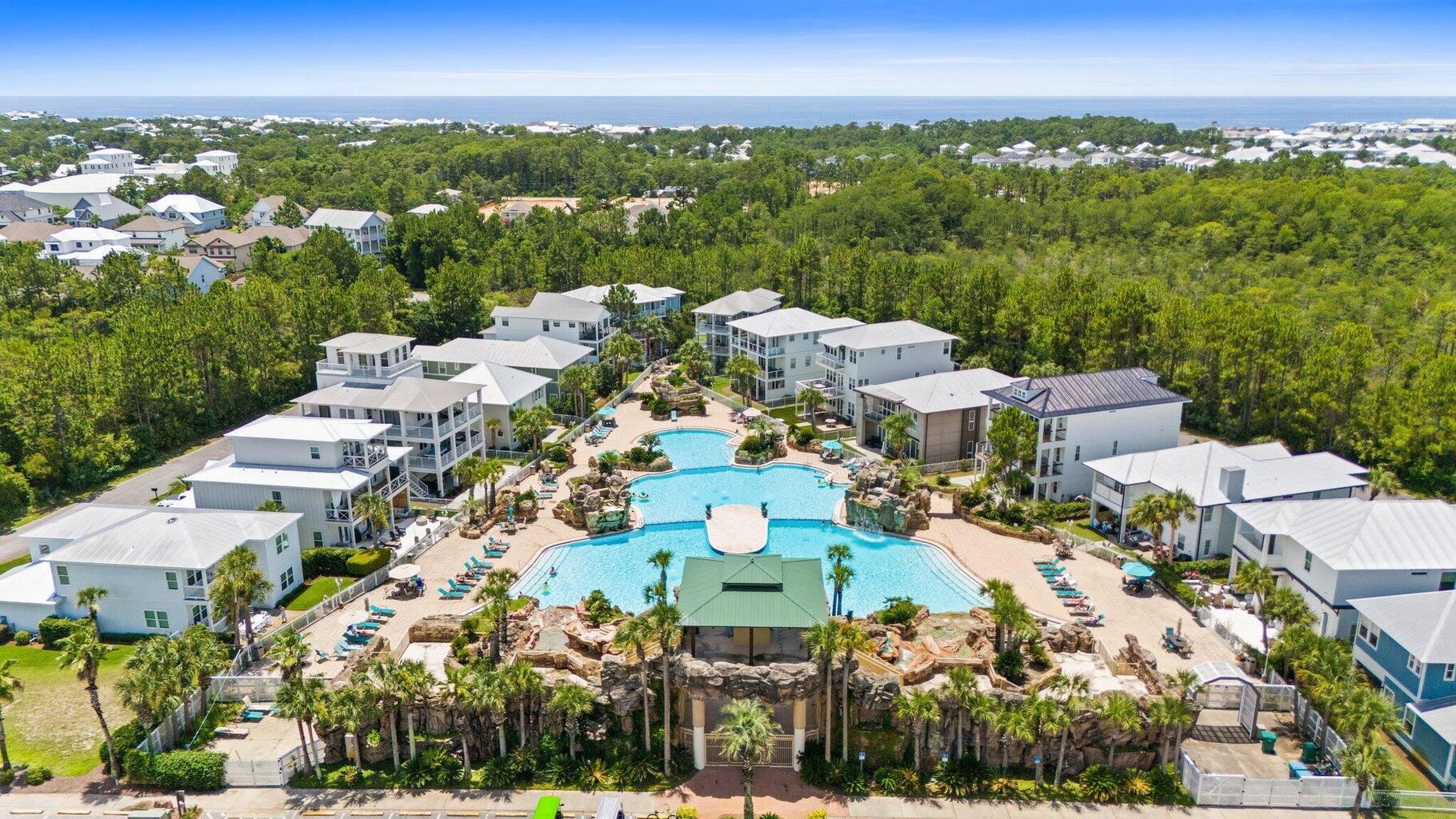 Aerial view of a residential community with a large pool, homes, trees, and the Gulf on the horizon.