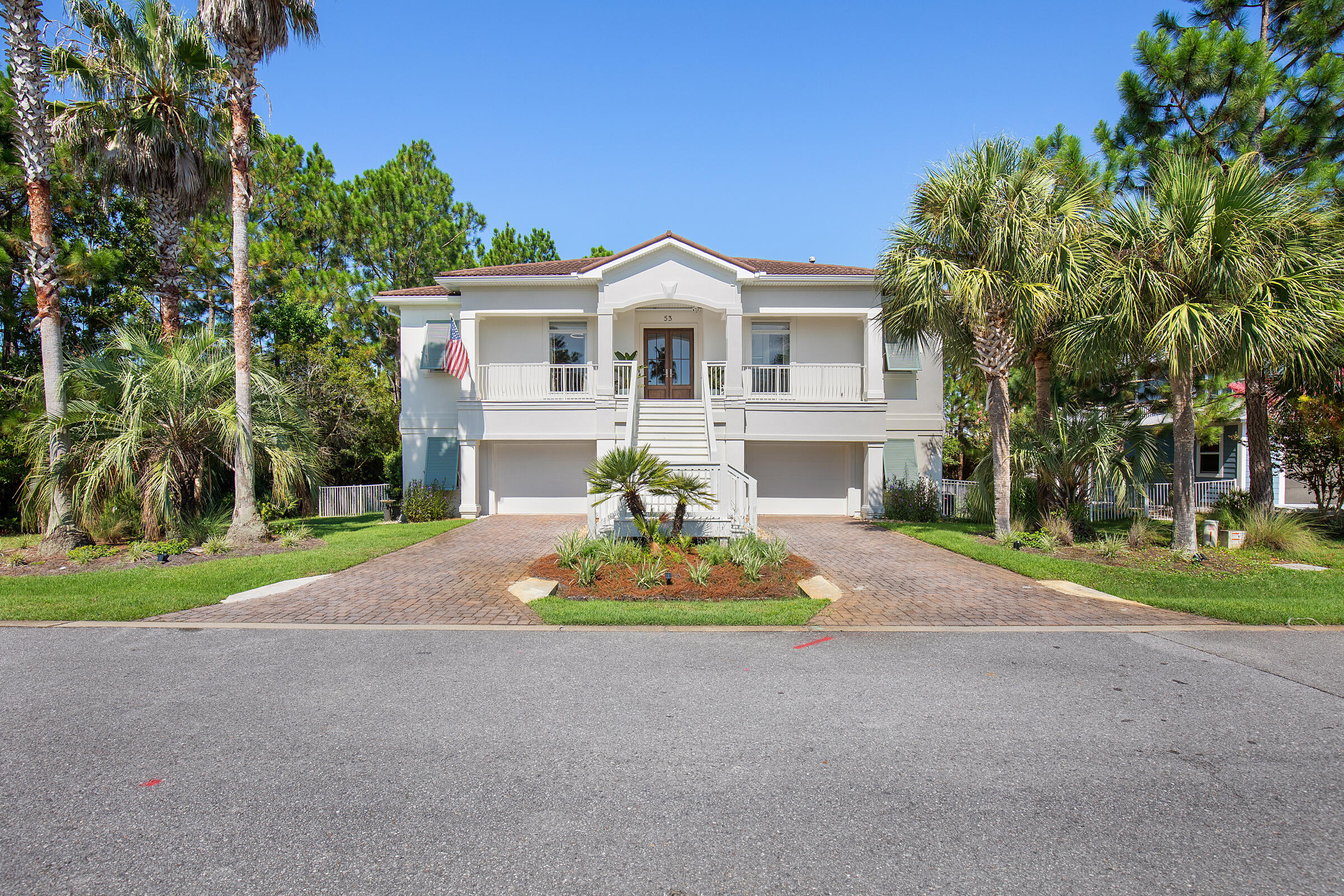 White raised house with a central staircase, paver driveway, and palm trees.