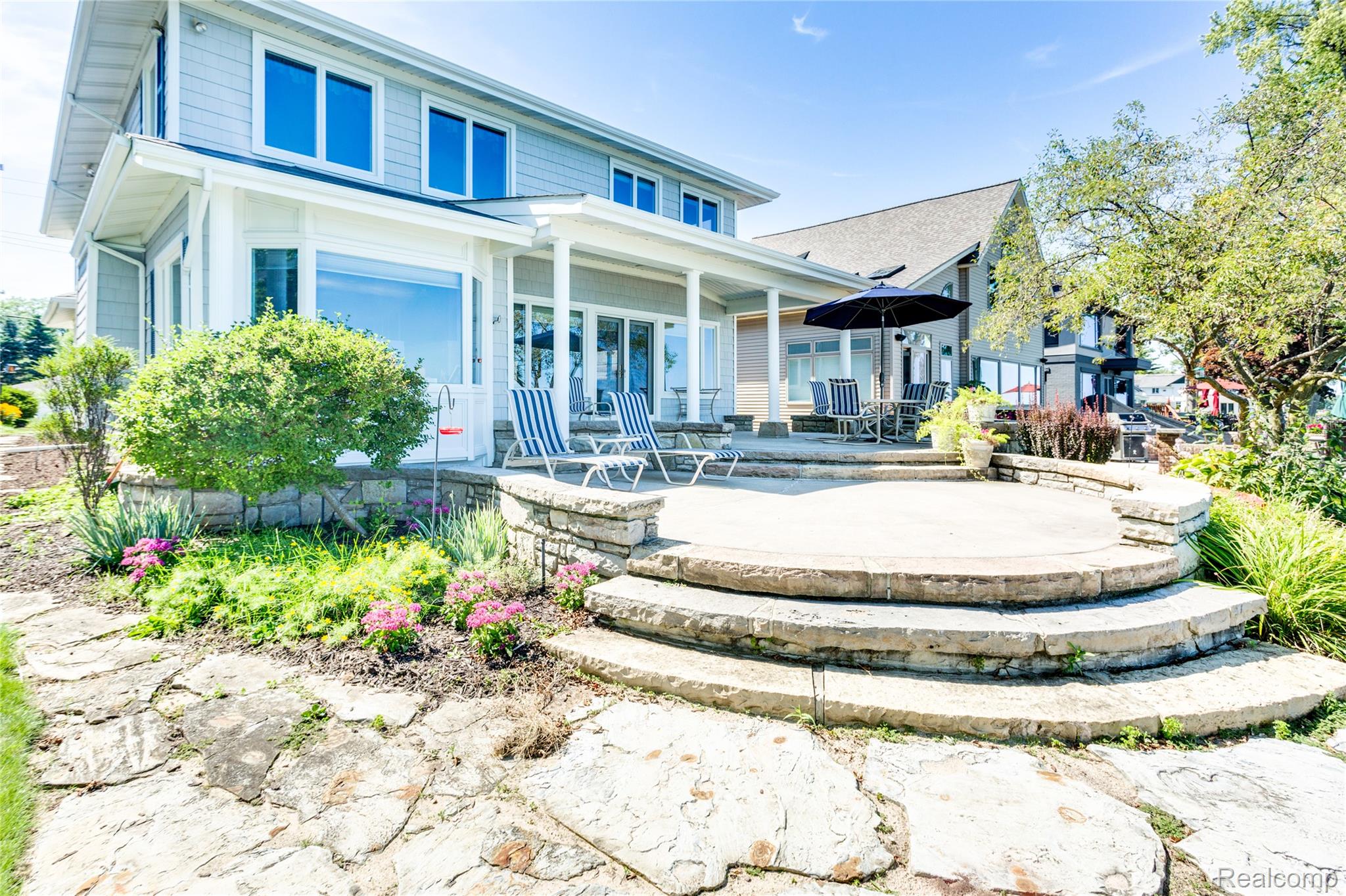Two-story home with a covered porch and curved stone patio furnished with striped lounge chairs.