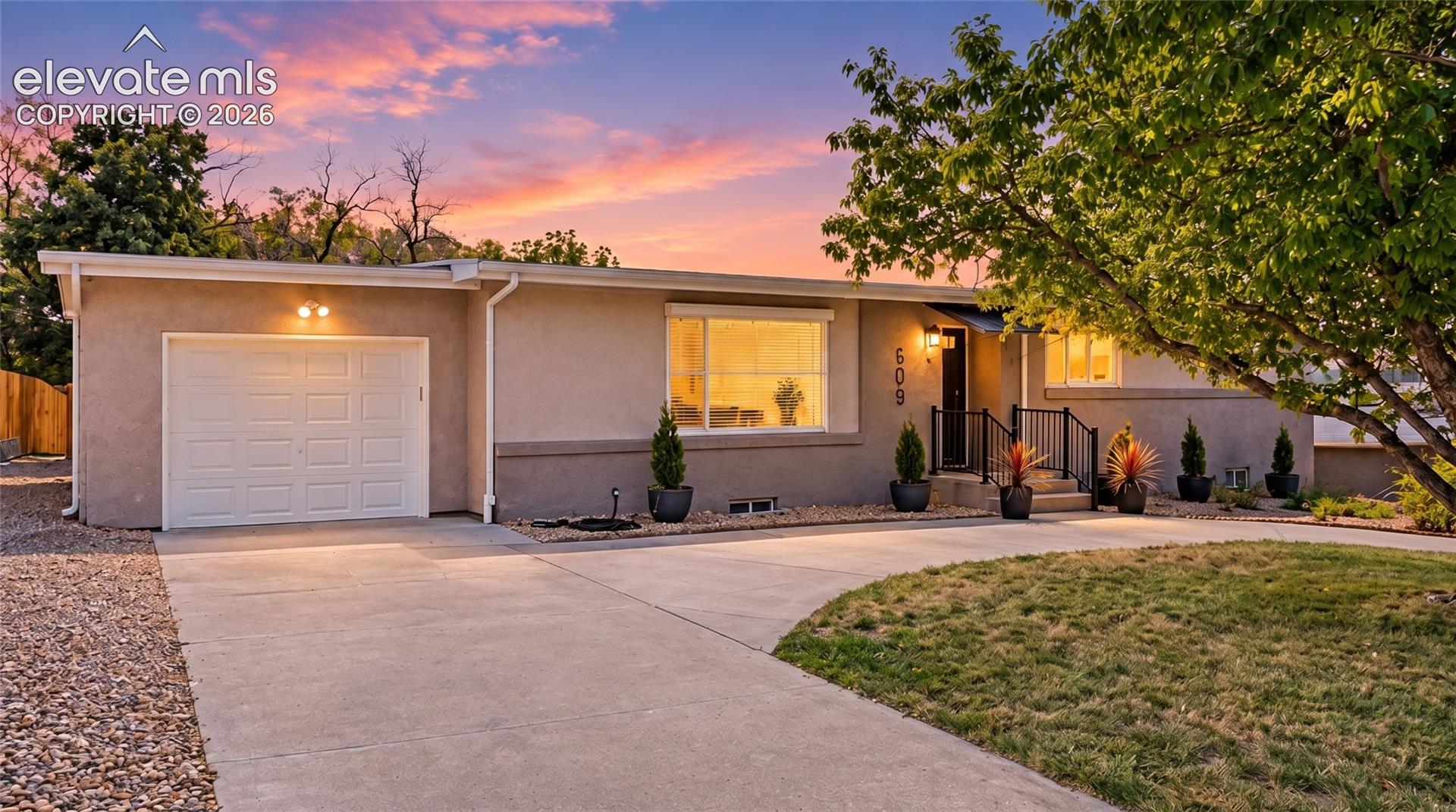 Single-story gray home with a white garage door and warmly lit windows beneath a pink dusk sky.
