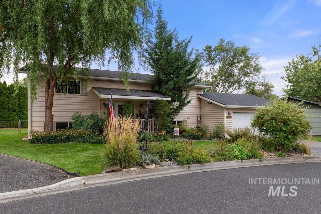 Beige split-level home with an attached garage, mature trees, and a landscaped front yard.