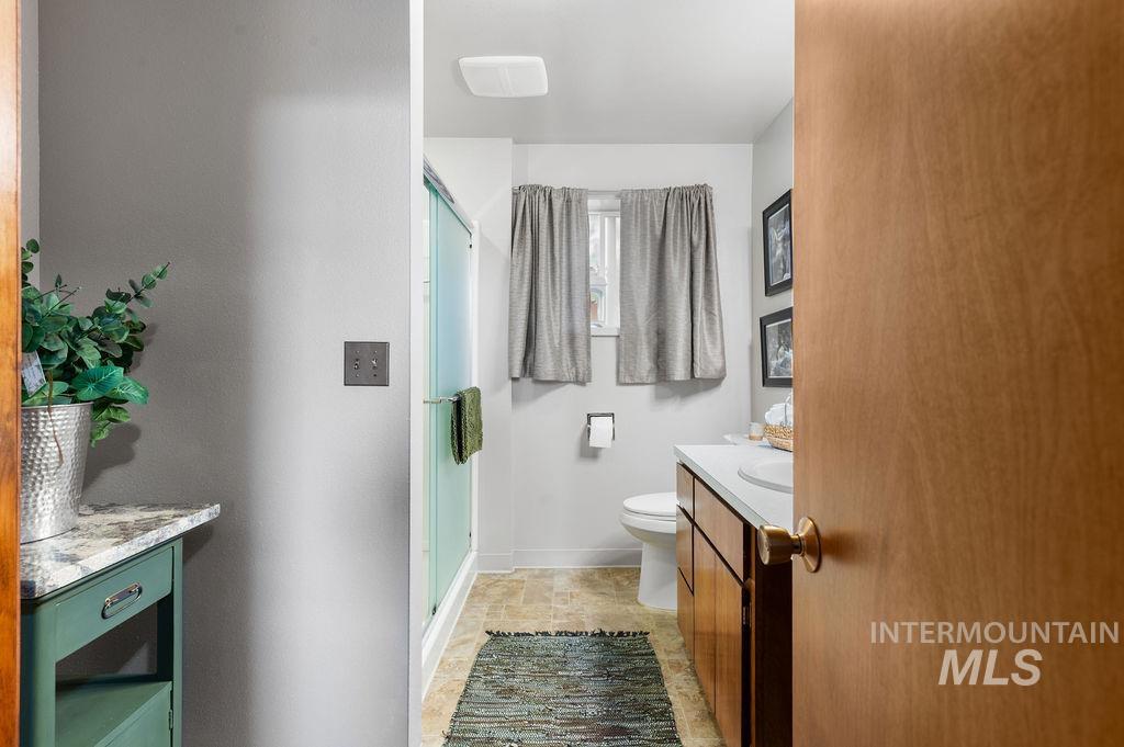Bathroom with a glass shower, wood vanity, and toilet beneath a curtained window.
