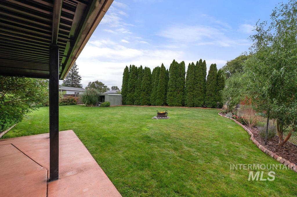 Backyard with a covered patio, broad lawn, central fire pit, and tall evergreen privacy screen.