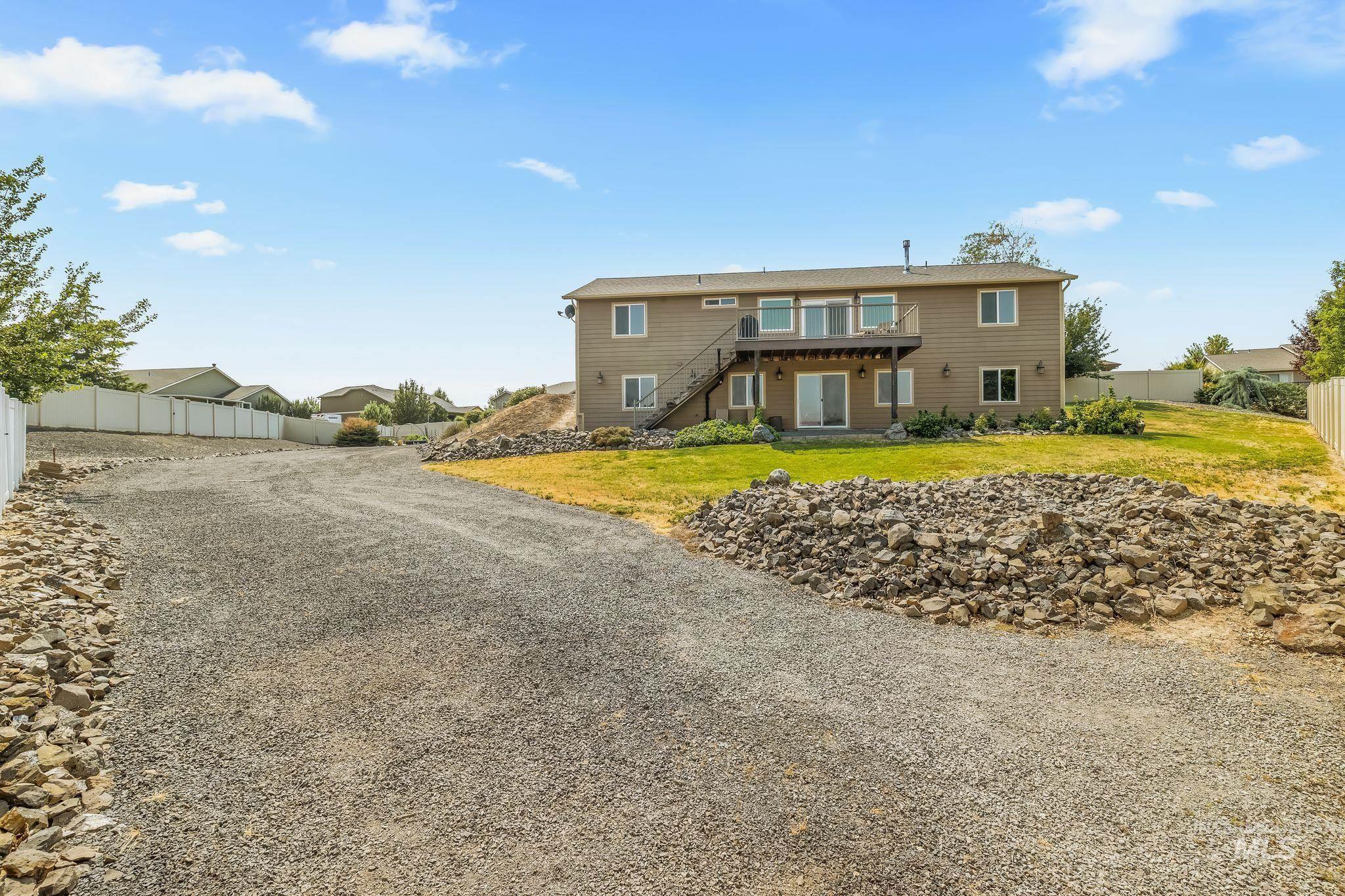 Two-story tan home with an elevated deck, exterior staircase, and broad gravel driveway.