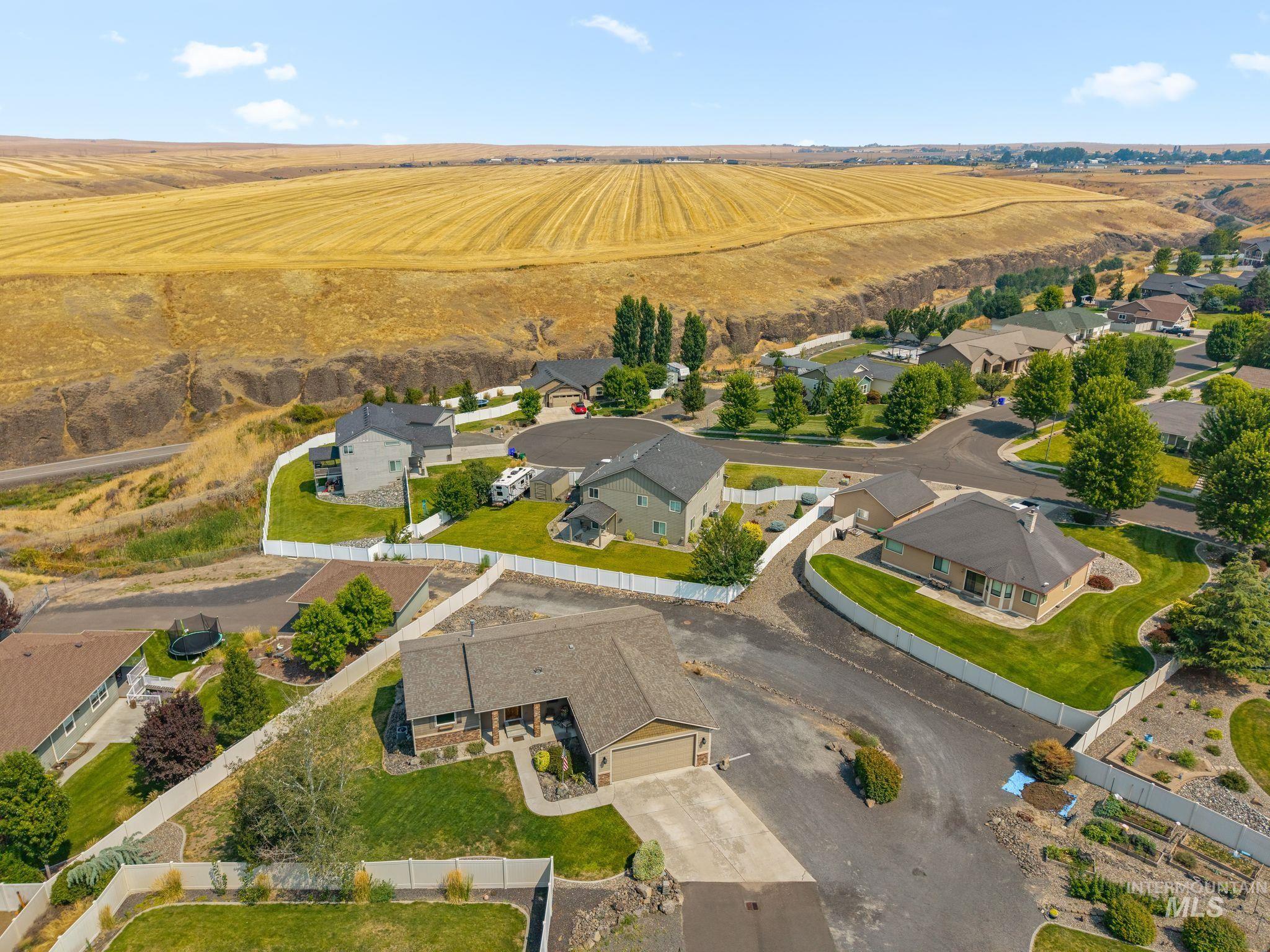 Aerial view of a home with a fenced lawn, neighboring houses, and golden fields beyond.