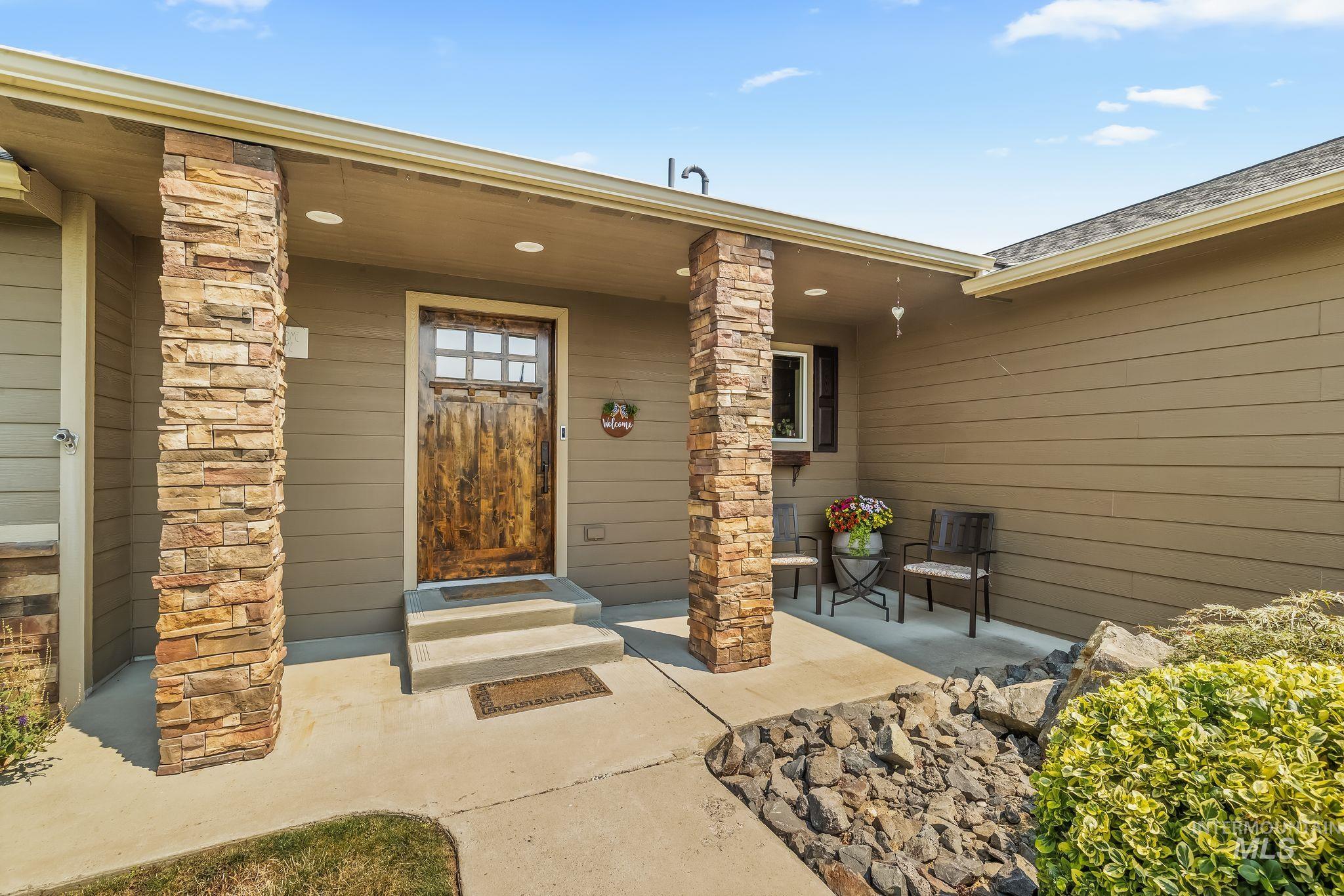 Covered home entry with stone columns, a rustic wood door, and porch chairs beside flowers.