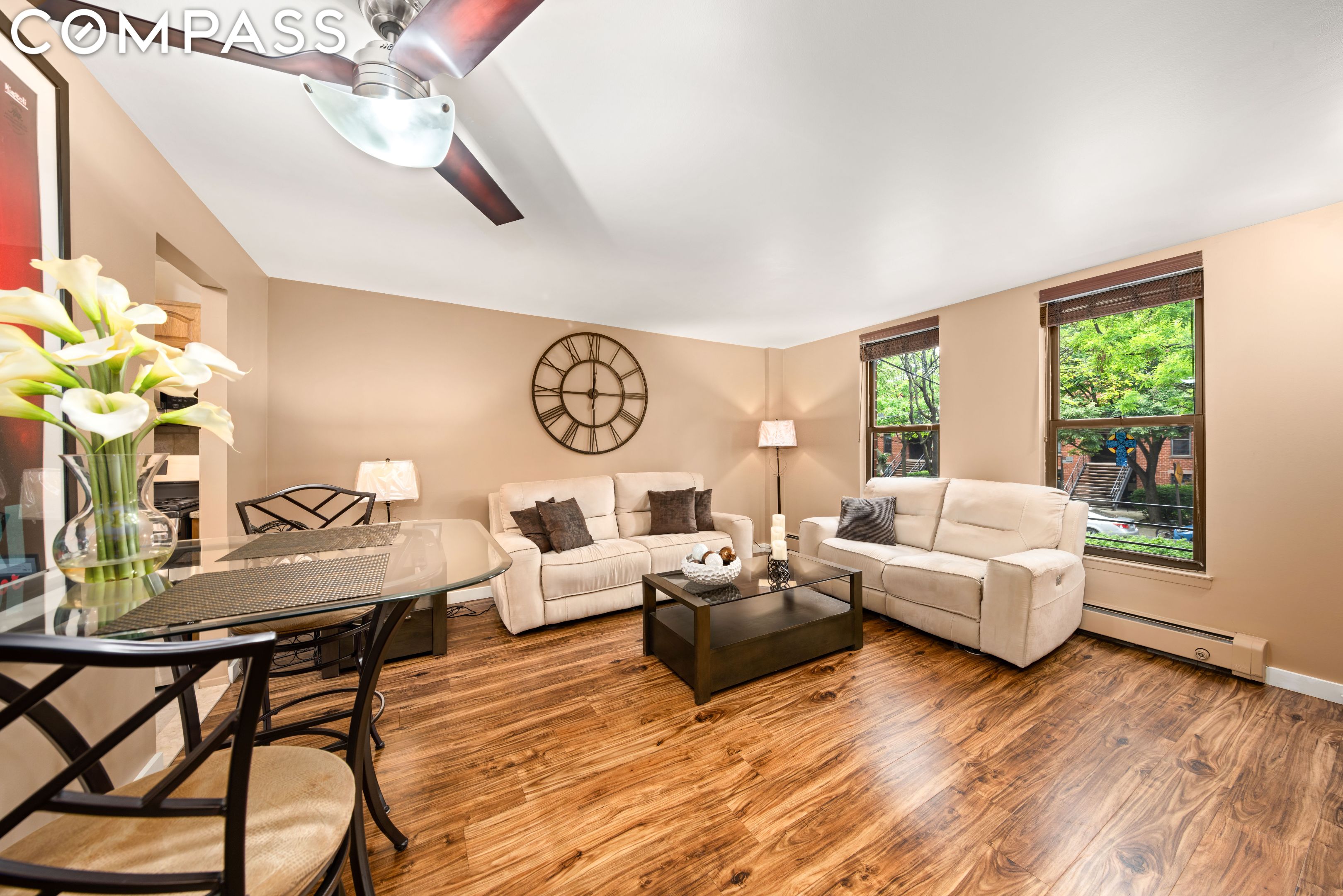 Staged living and dining room with cream sofas, wood floors, and windows overlooking leafy trees.