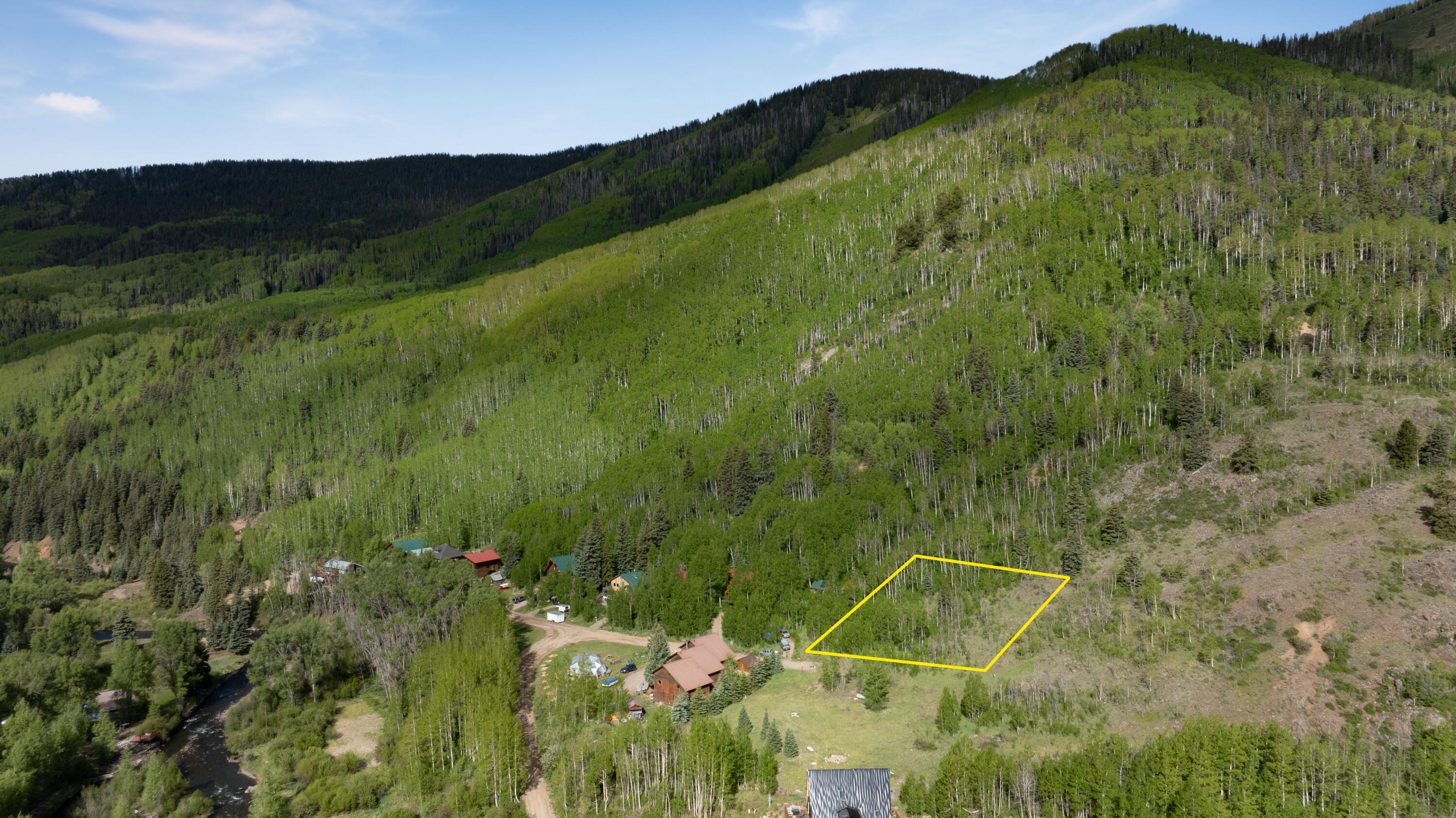 Aerial view of a yellow-outlined wooded lot beside homes, a river, and a forested mountain.