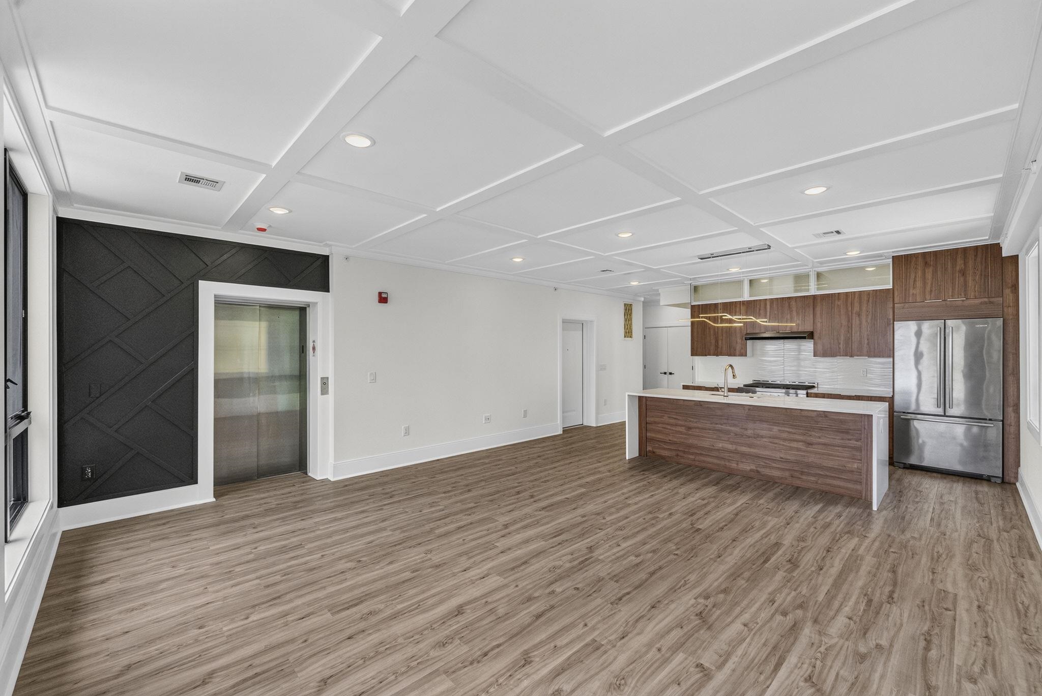 Unfurnished open-plan living area with a wood-front kitchen island, stainless steel appliances, and geometric black accent wall.