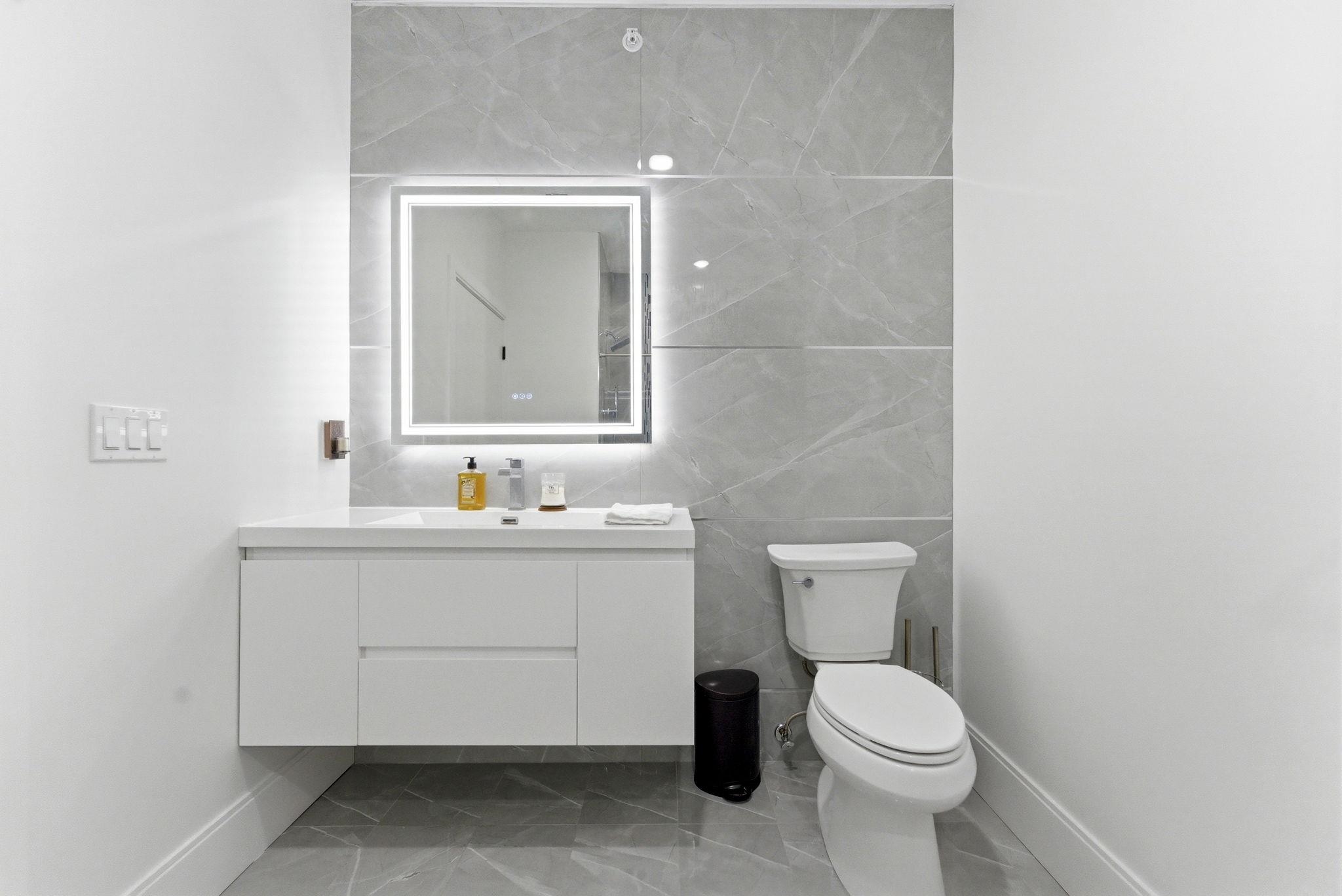 Modern bathroom with a floating white vanity, illuminated mirror, gray tile, and toilet.
