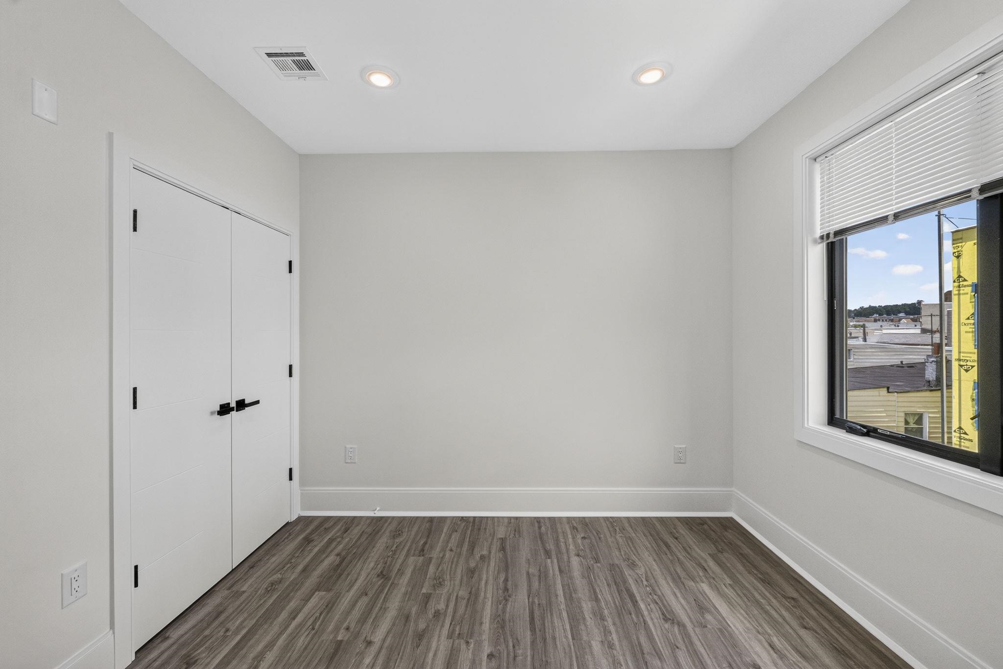 Empty room with dark wood-look flooring, double closet doors, and a large window with blinds.