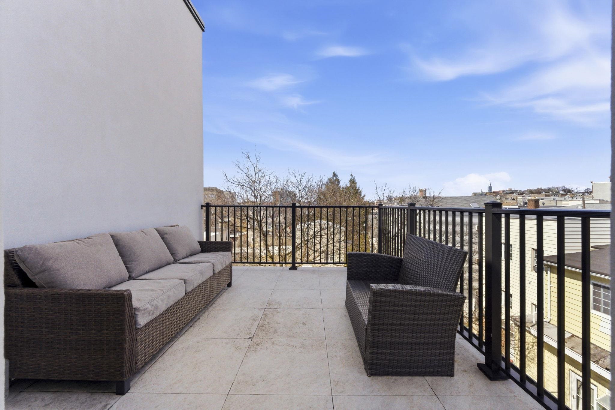 Furnished balcony with woven seating, tiled floor, and views of nearby rooftops.