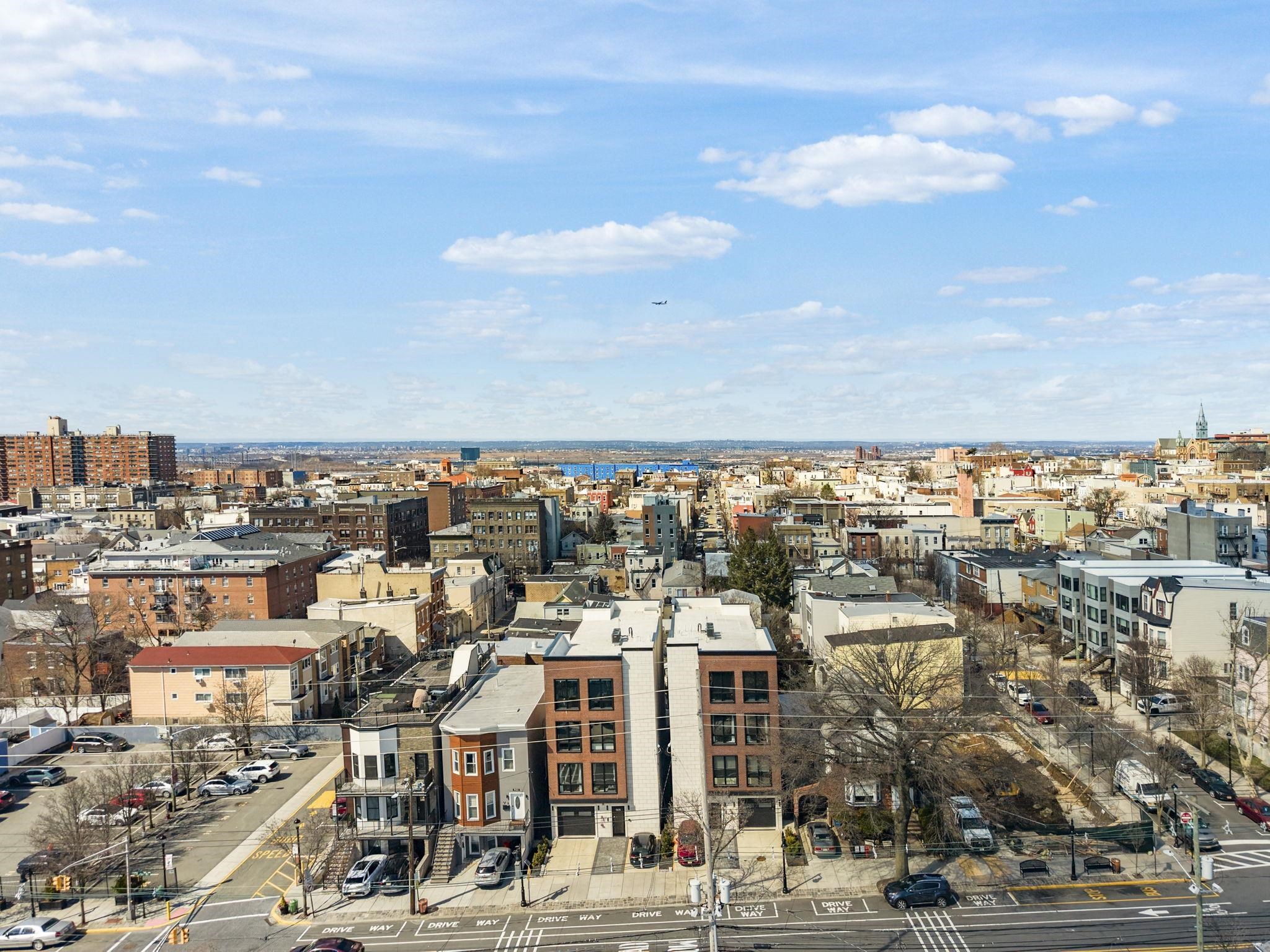 Aerial view over dense city buildings, with a church spire and distant skyline beneath a broad blue sky.